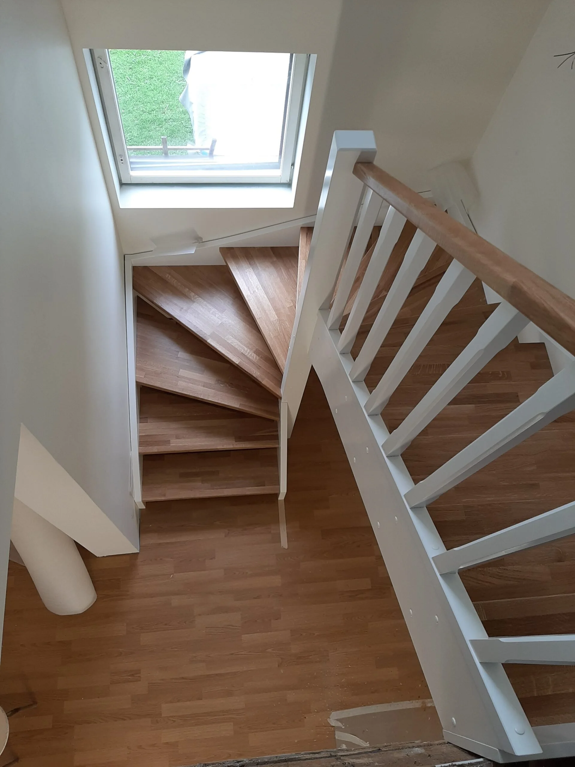 View of a wooden staircase with white railing from above, next to a window showing an outdoor area with grass and a person standing outside.