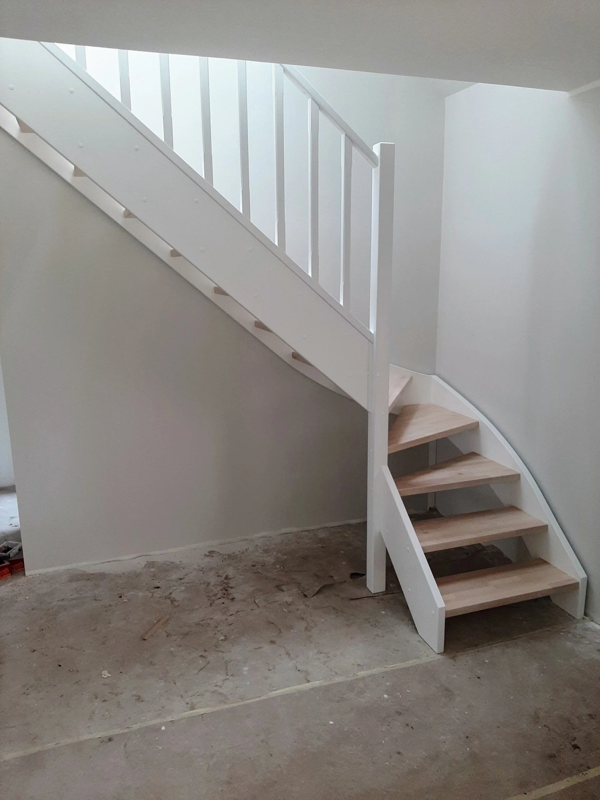 Interior view of a partially constructed staircase with wooden steps, white railings, and a wall, on an unfinished concrete floor.