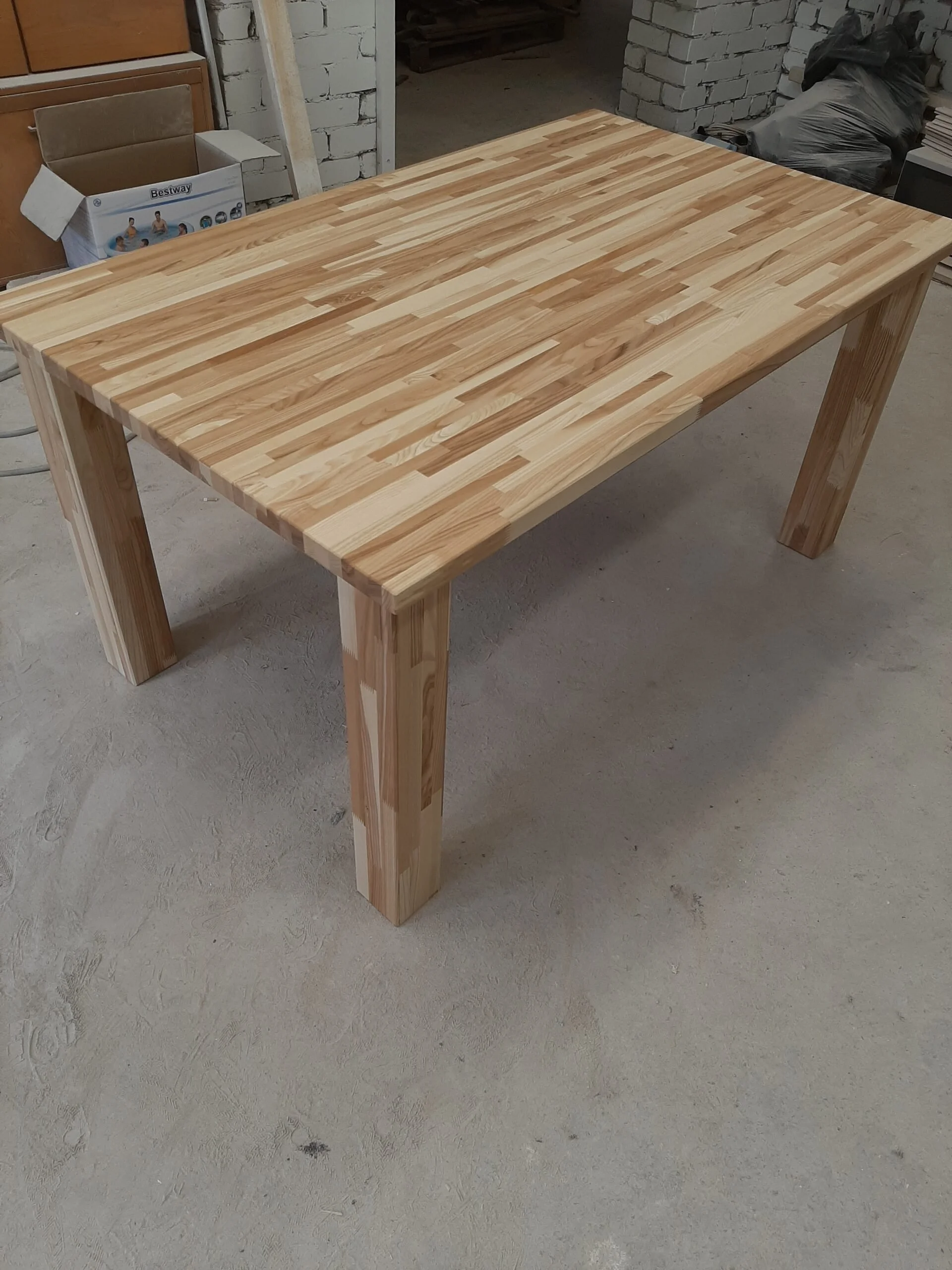 A wooden table with a striped pattern on the tabletop, constructed from light-colored wood planks, in a workshop setting.
