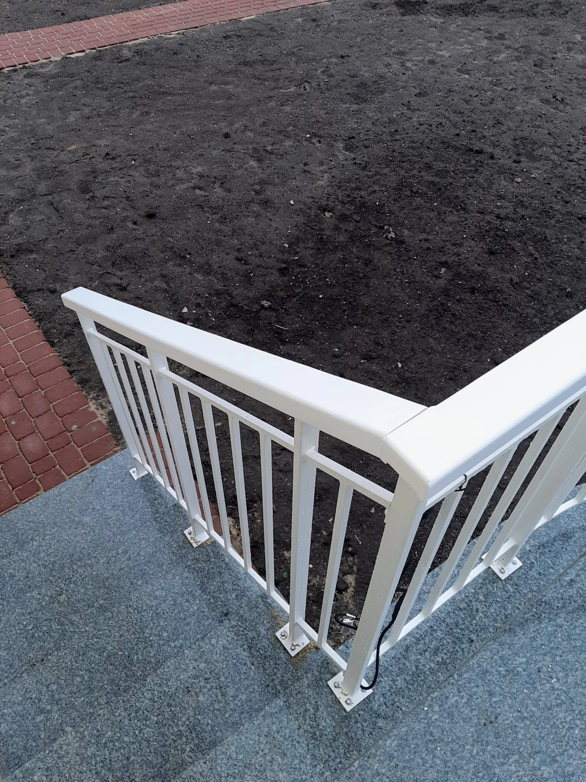 White metal railing around a small, enclosed area with dark dirt ground and a brick sidewalk nearby.