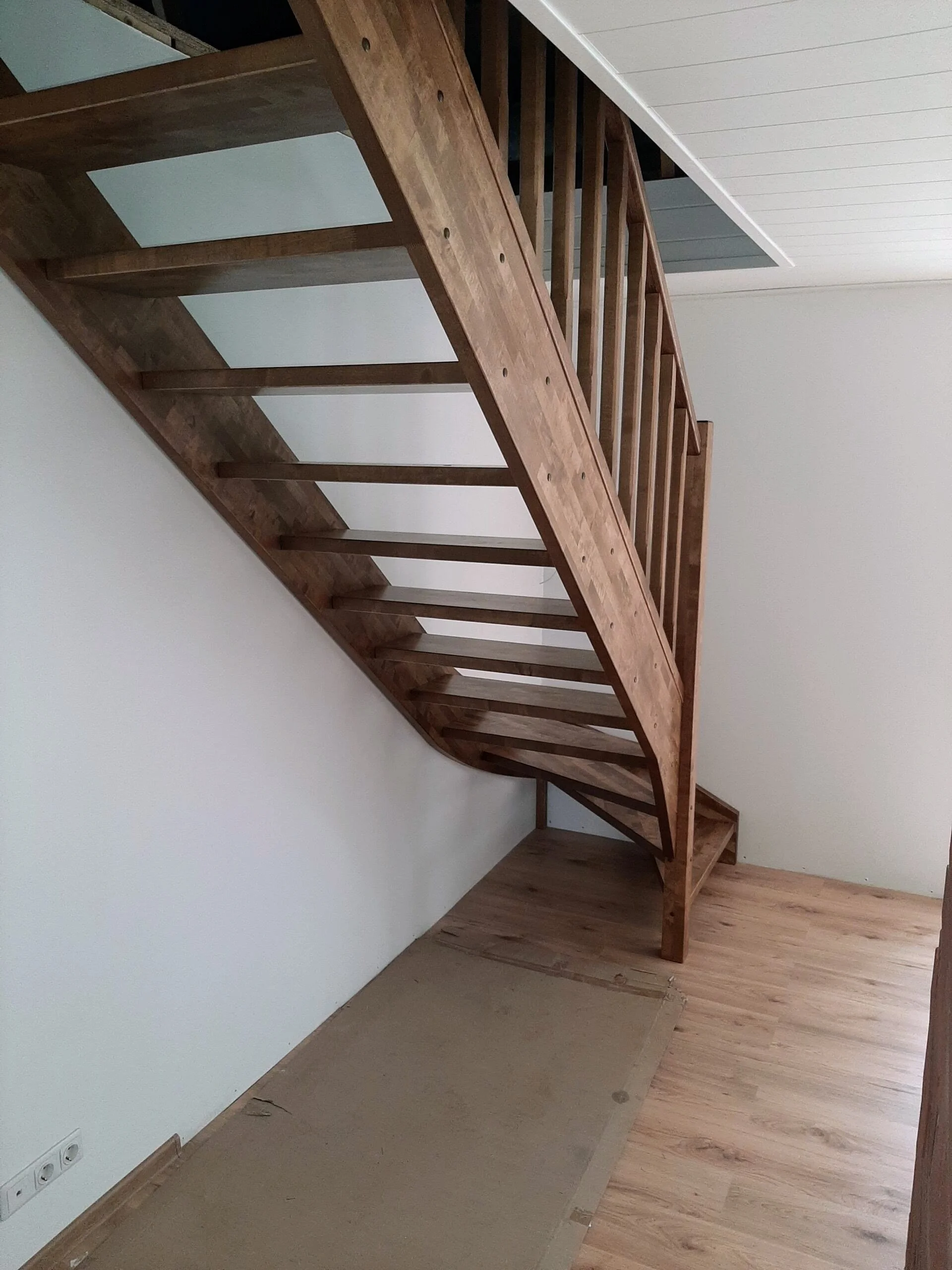 Wooden staircase with a curved design leading to an upper floor, next to a white wall with electrical outlets at the bottom.