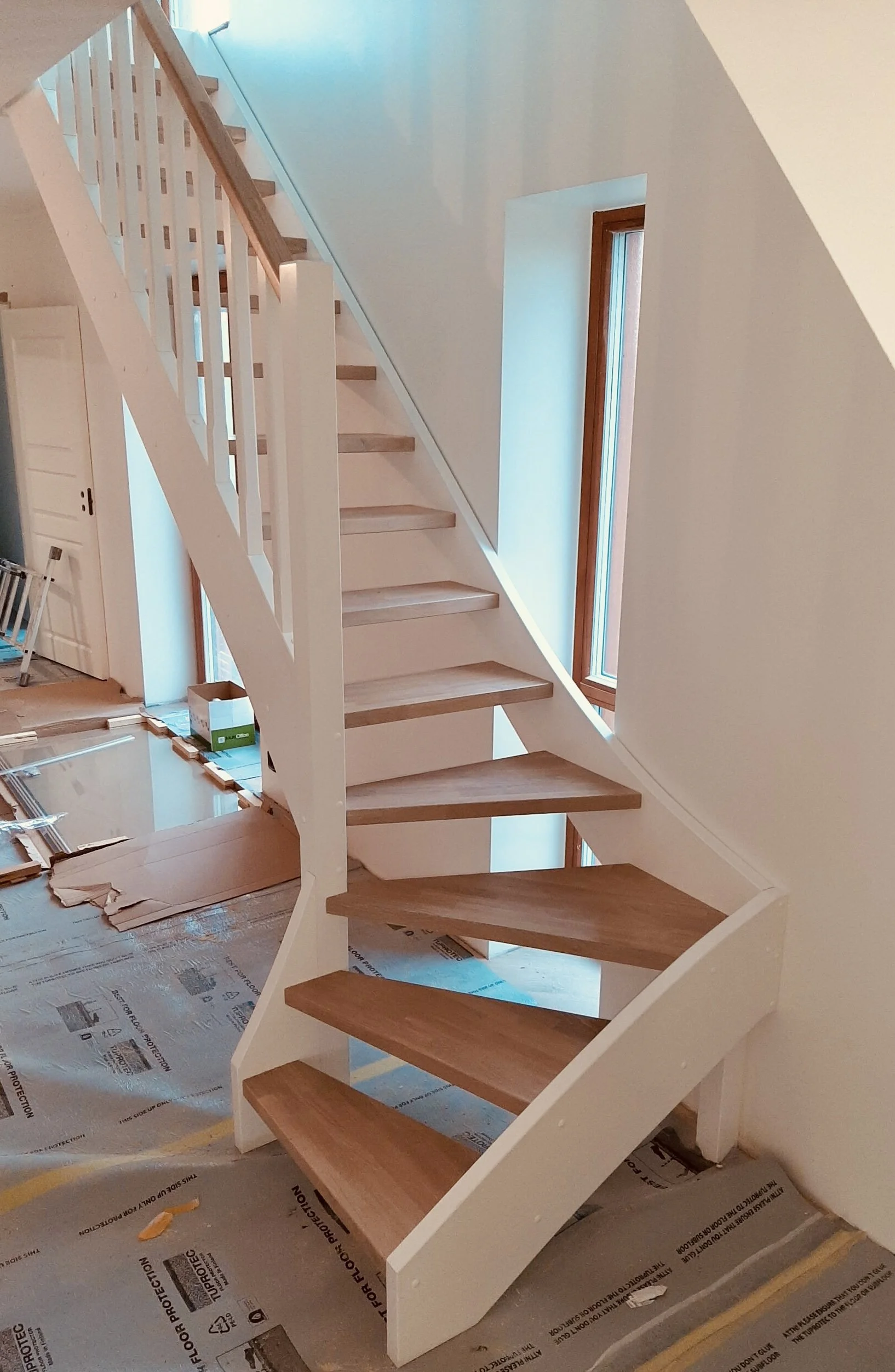 A modern staircase with wooden steps and white safety railing inside a house under construction, with cardboard on the floor and large windows letting in natural light.