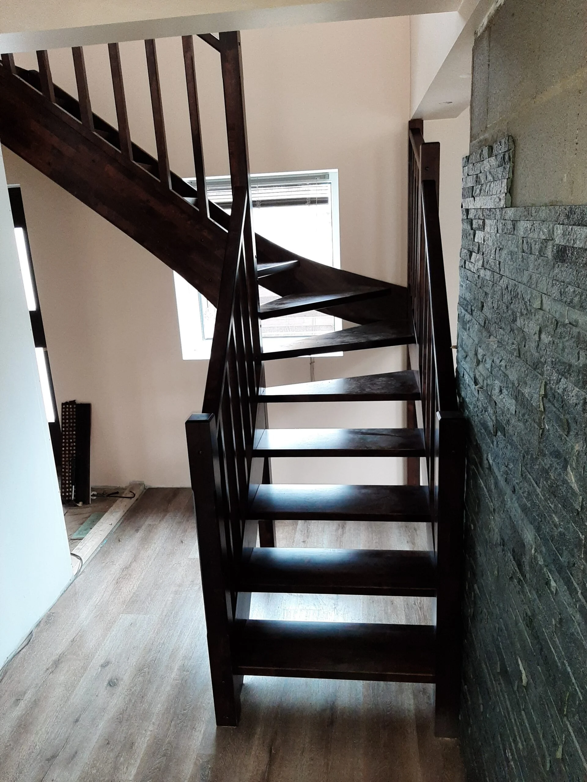 Interior of a house showing dark wooden stairs with a simple railing, next to a stone wall. There is a window providing natural light and wooden flooring.