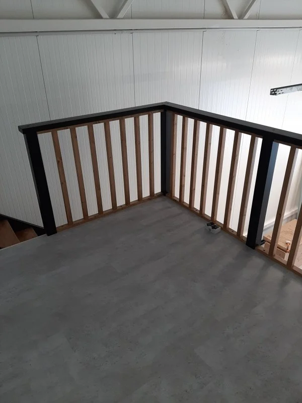 Interior view of a balcony or loft area with a concrete floor, wooden railing with vertical slats, and white wall panels in the background.