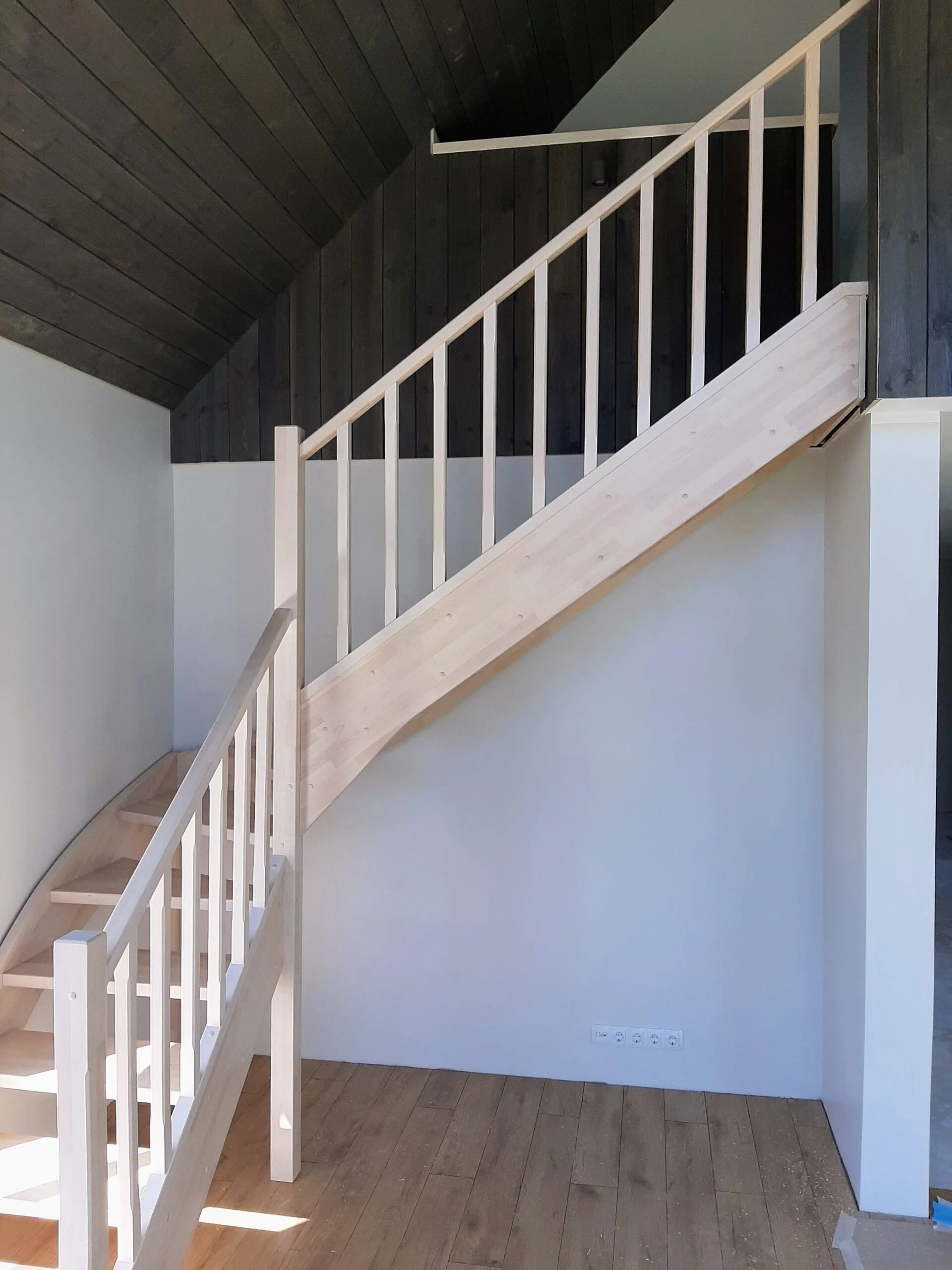 Interior view of a wooden staircase with new light wood steps and white railing, leading to a dark wood ceiling with a sloped design.