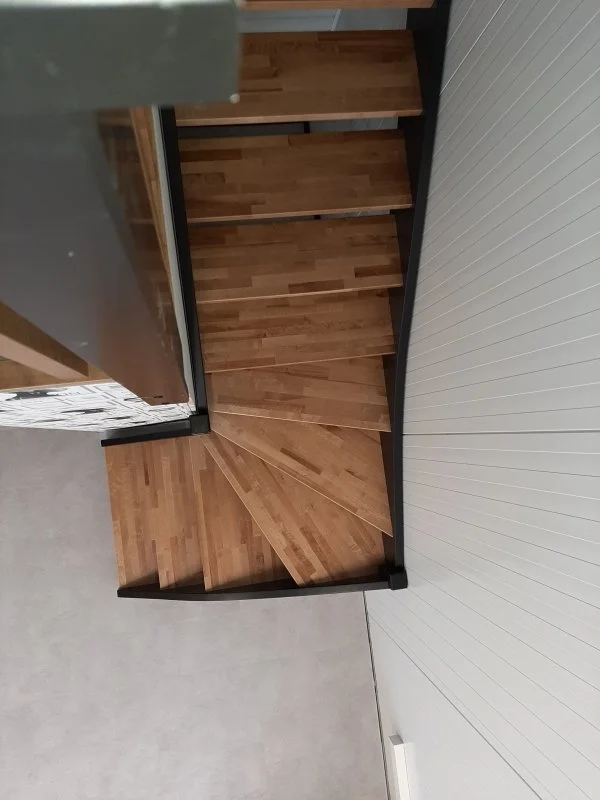 Wooden spiral staircase with black metal railing viewed from below, inside a room with white paneled wall.