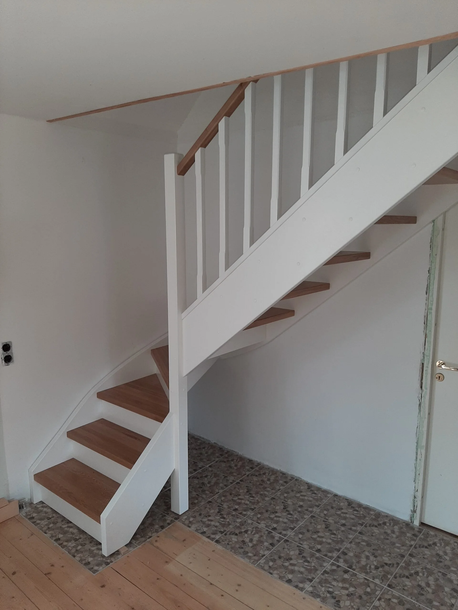 White staircase with wooden steps and wooden handrail inside a house, with exposed wall on the right and tiled floor underneath.