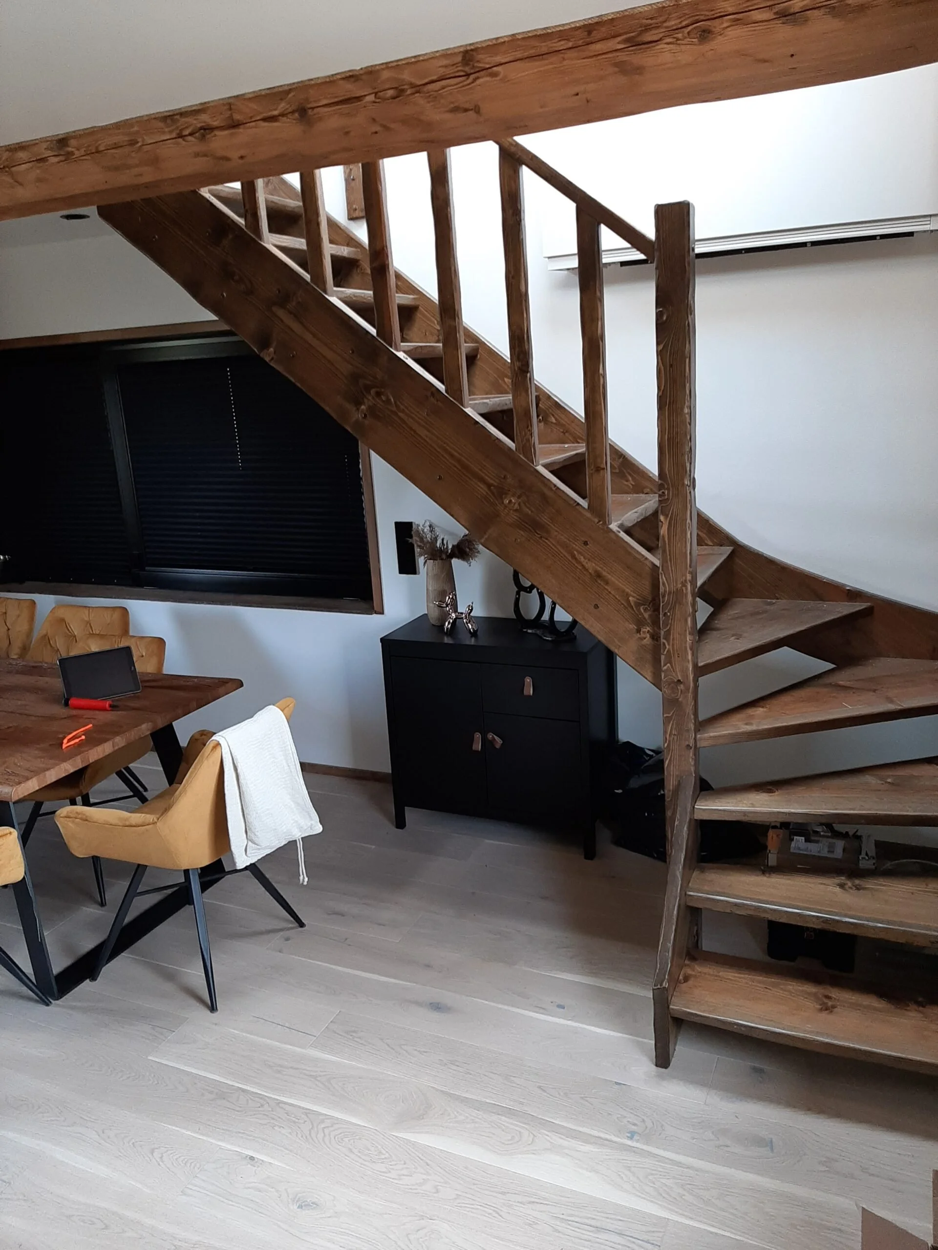 Interior view of a room showing a wooden staircase, a black cabinet with decorative items, a window with closed blinds, a wooden dining table with chairs, a towel on one chair, and a tablet and pen on the table.