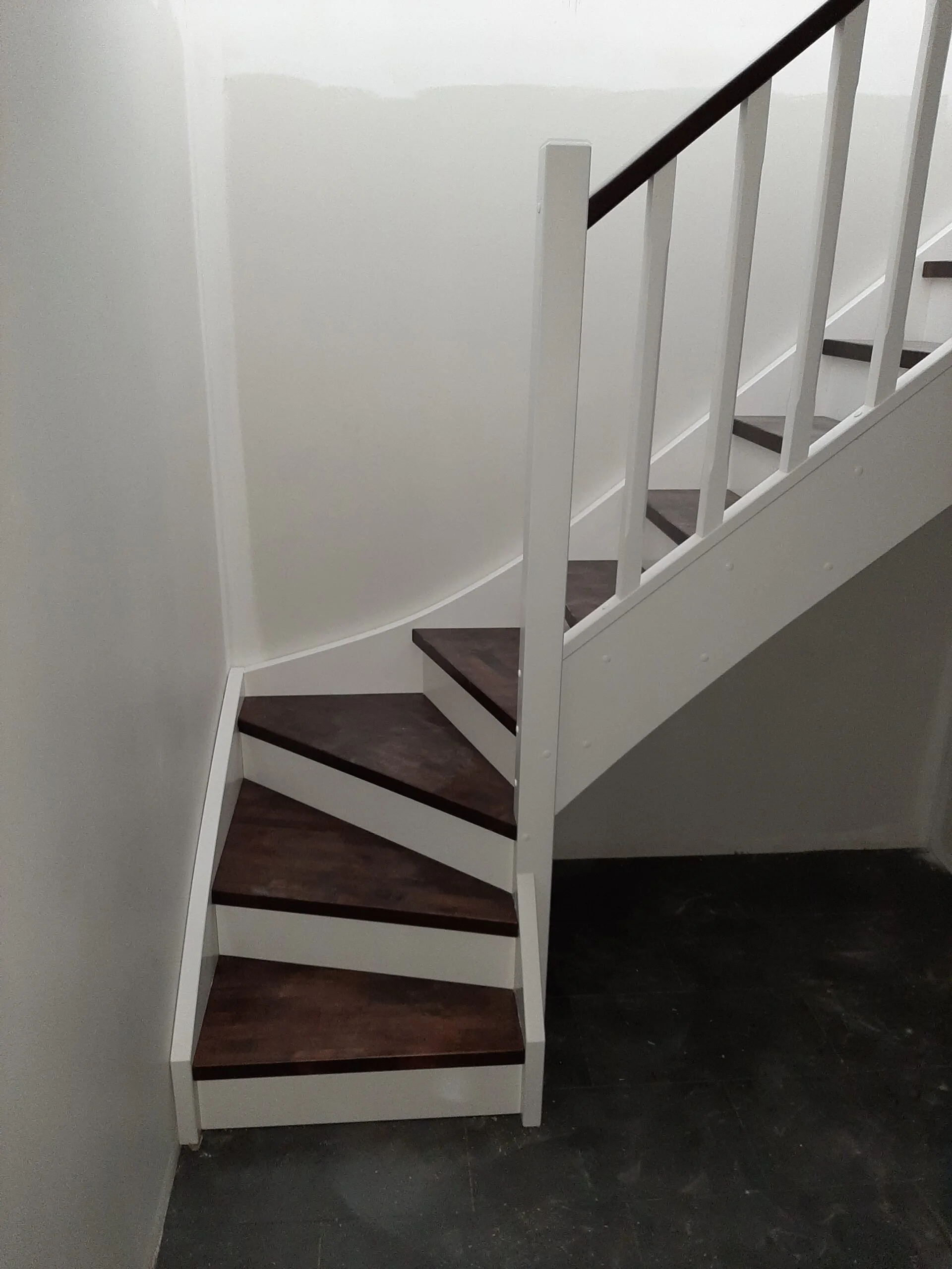 A staircase with dark wood steps, white risers, and a white railing with dark handrail, ascending along a white wall.