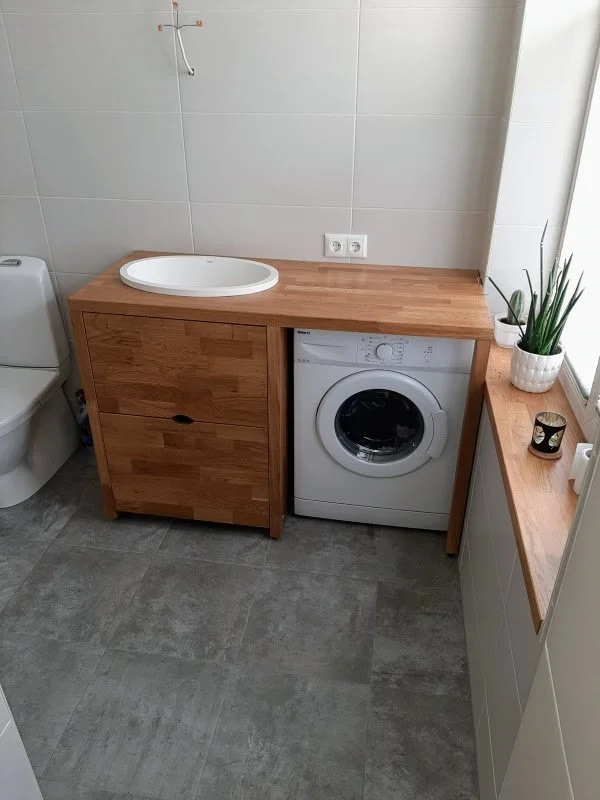 Laundry room with washing machine integrated into wooden countertop, a small sink on top, a toilet to the left, potted plants on the right window sill, and gray tiled flooring.