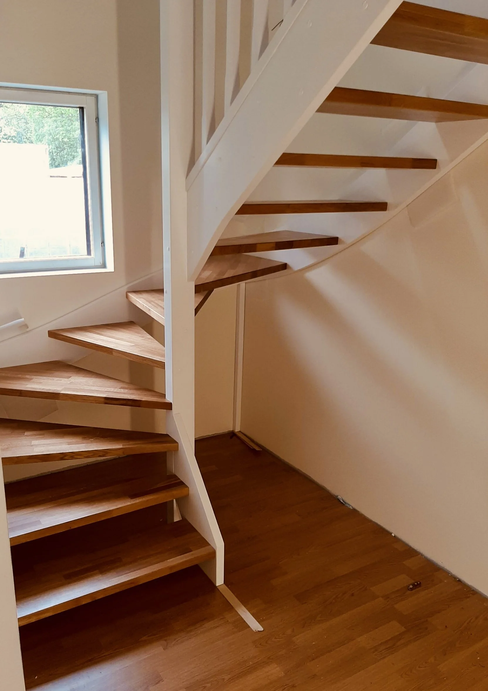 Interior view of a wooden staircase spiraling upward next to a window in a home.