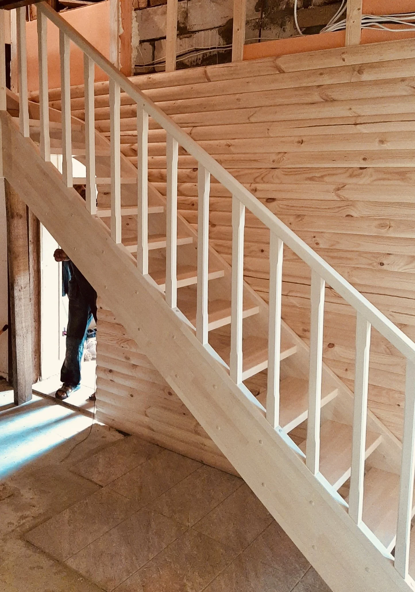 Wood staircase under construction inside a building, with partially finished wooden walls and flooring, and a person working near the door.