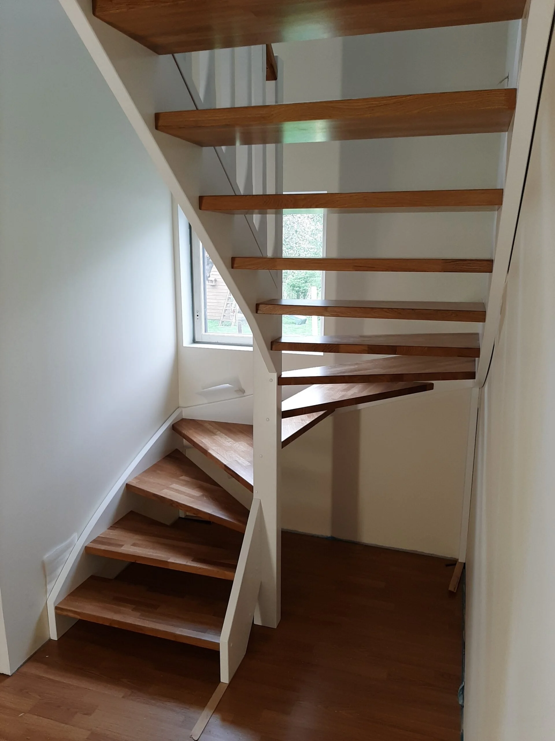 Interior view of a wooden staircase with open risers and a curved design, adjacent to a window with a view of a garden.