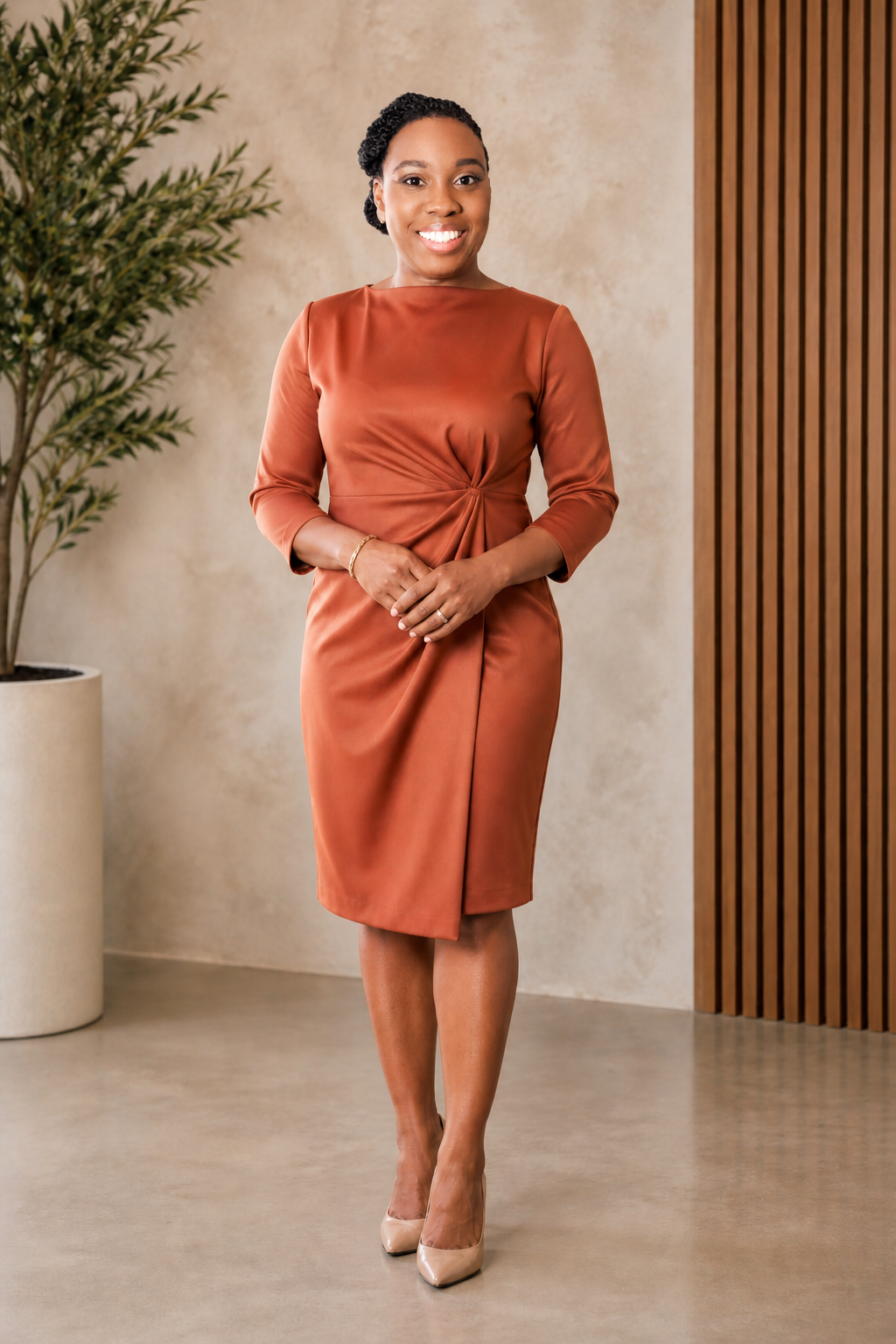 A woman in an elegant rust-colored dress standing indoors with a potted plant on her left and wooden paneling on her right, smiling at the camera.