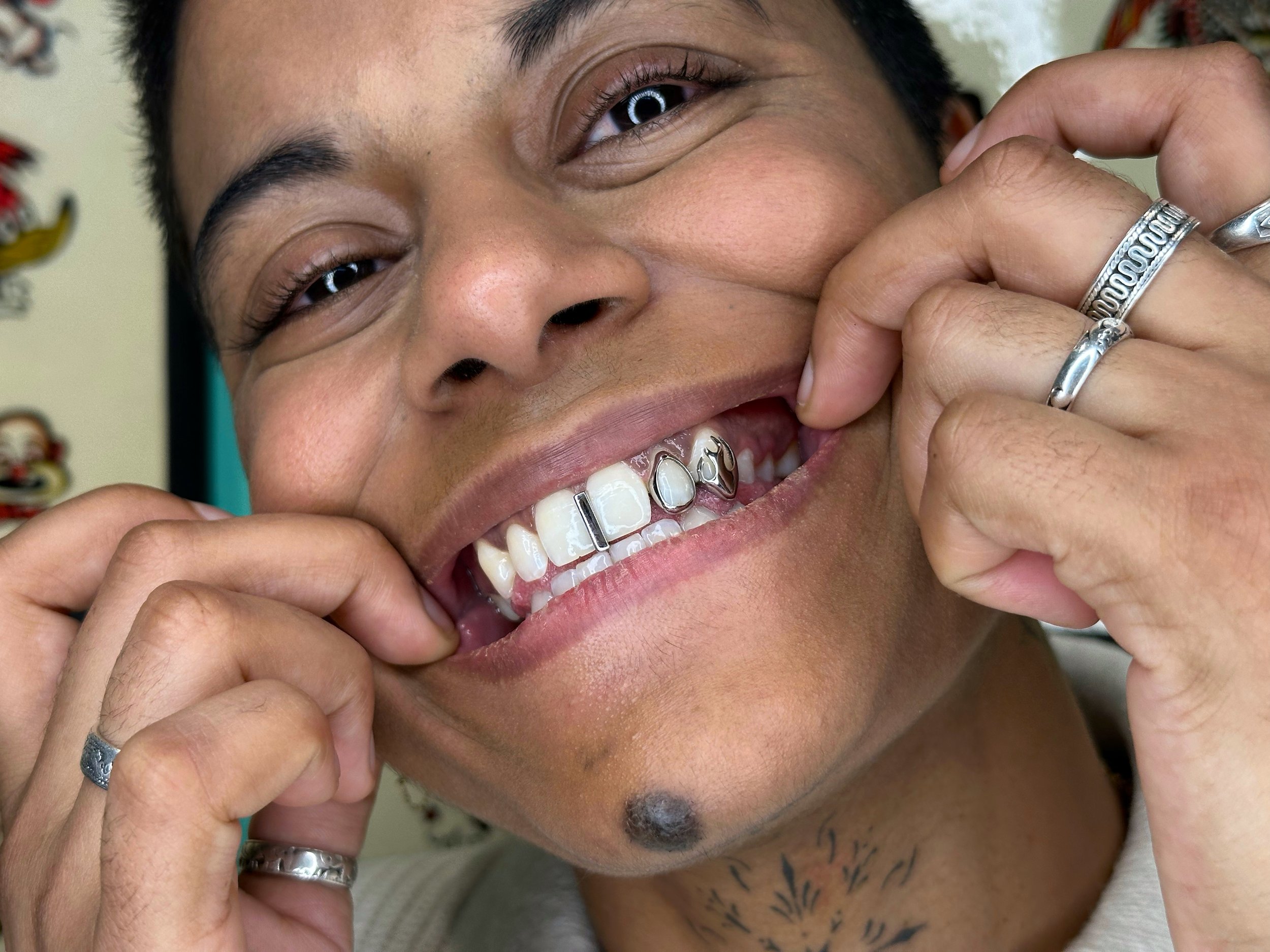 Close-up of a person smiling and pulling their lips to show their teeth and jewelry, with background items.