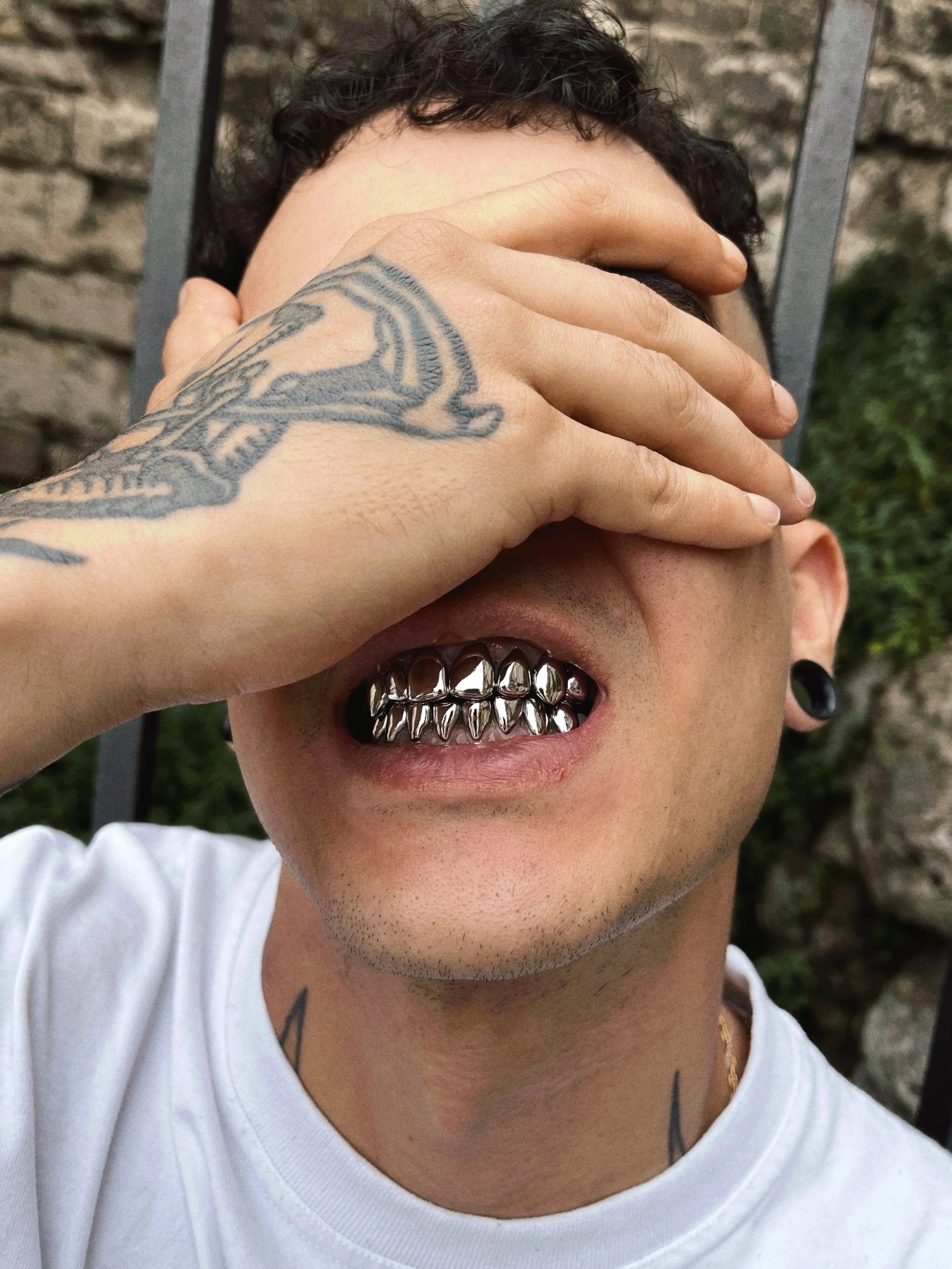 Person with dark curly hair, tattoos, and gauges, covering one eye with one hand, showing metal grill teeth, wearing a white shirt, outdoors.