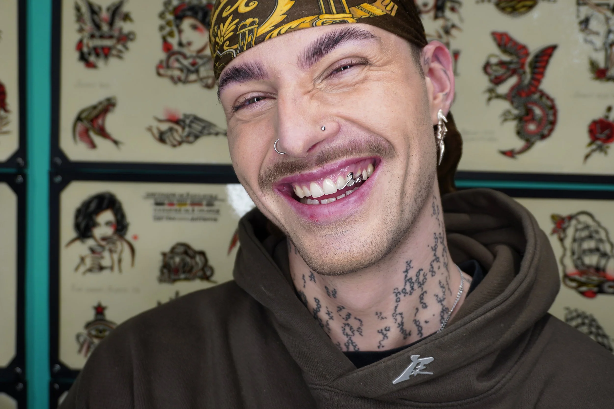 Close-up of a smiling man with tattoos, piercings, and a bandana, standing in front of tattoo flash art.