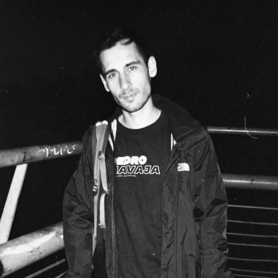 A man standing outdoors at night on a bridge or railing, wearing a jacket and a T-shirt, with a backpack, looking at the camera.