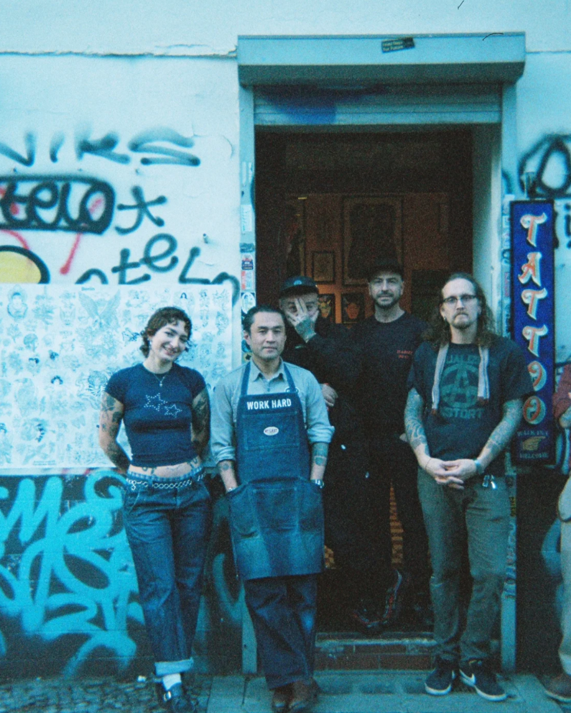 Group of five people standing in front of a graffiti-covered wall and restaurant entrance, some with tattoos, dressed casually.