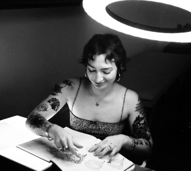 A smiling woman with tattoos on her arms, wearing a sleeveless top, reading a book at a table in a dimly lit room.