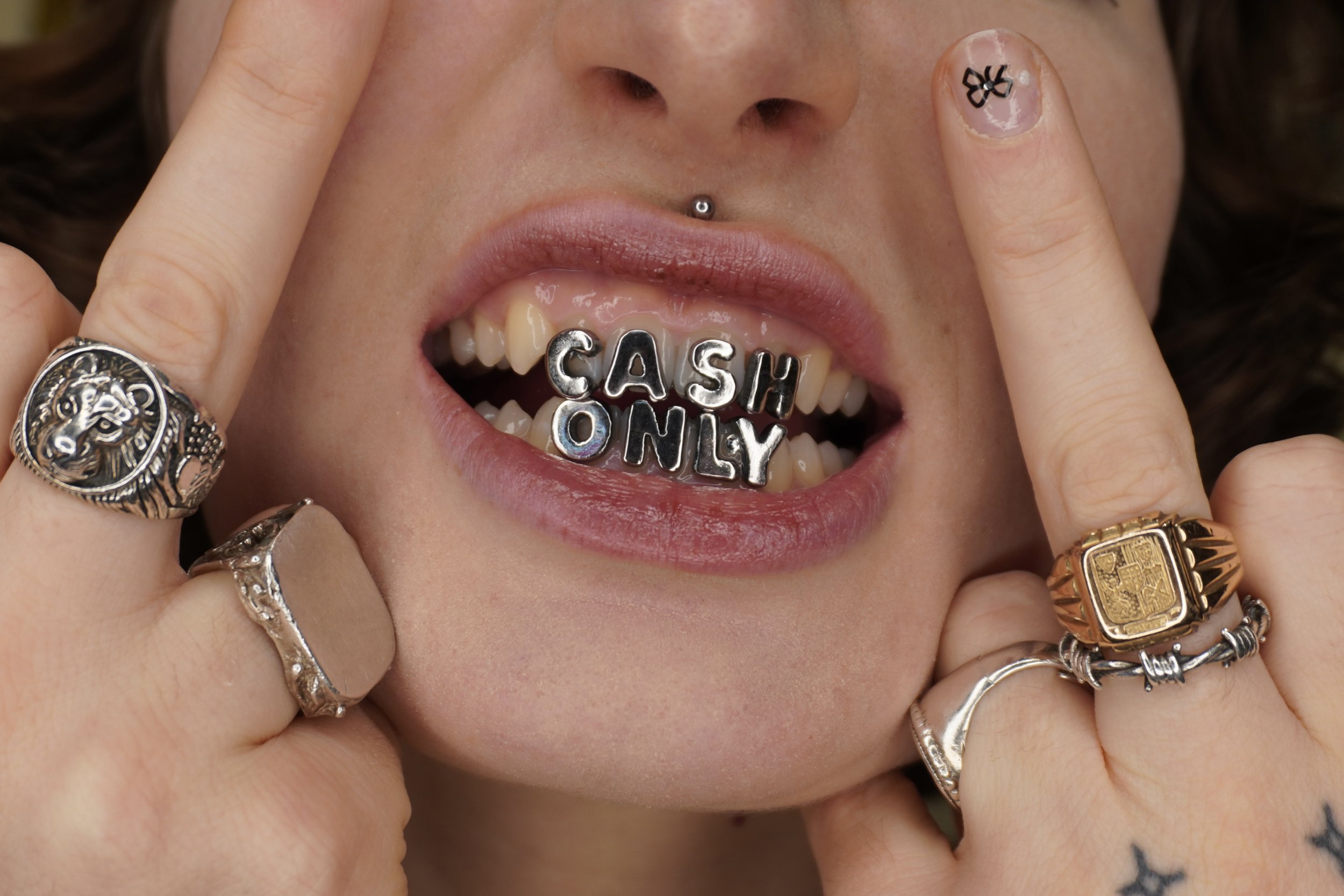 Close-up of a person with jewelry and finger tattoos holding their lips open, revealing a tongue with letter and word beads spelling 'CASH ONLY' in their mouth.