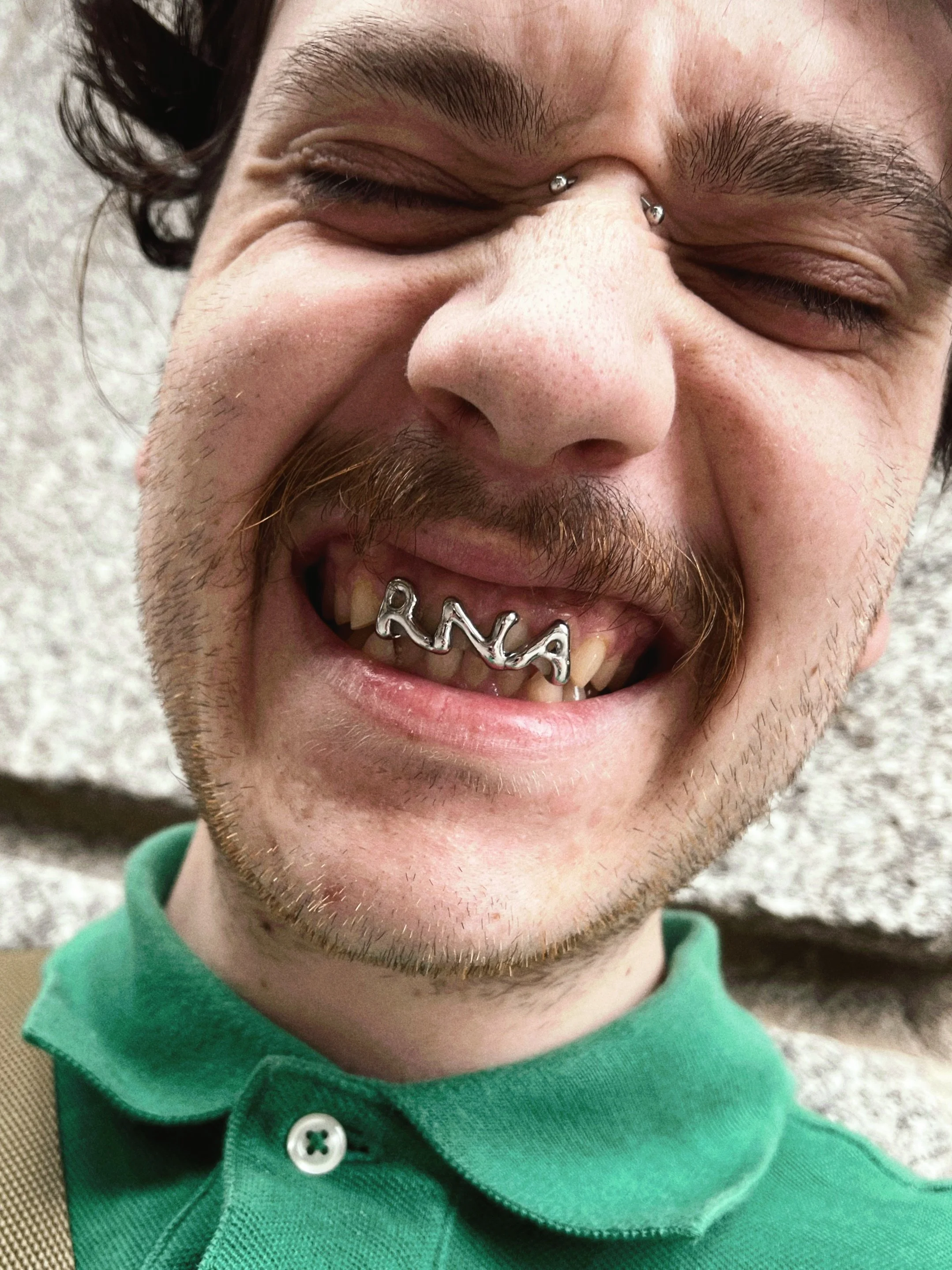 A young man with a beard smiling with his eyes closed, wearing a green polo shirt and a silver nose ring, showing a silver jewelry piece on his lower lip with the name 'RANA'.