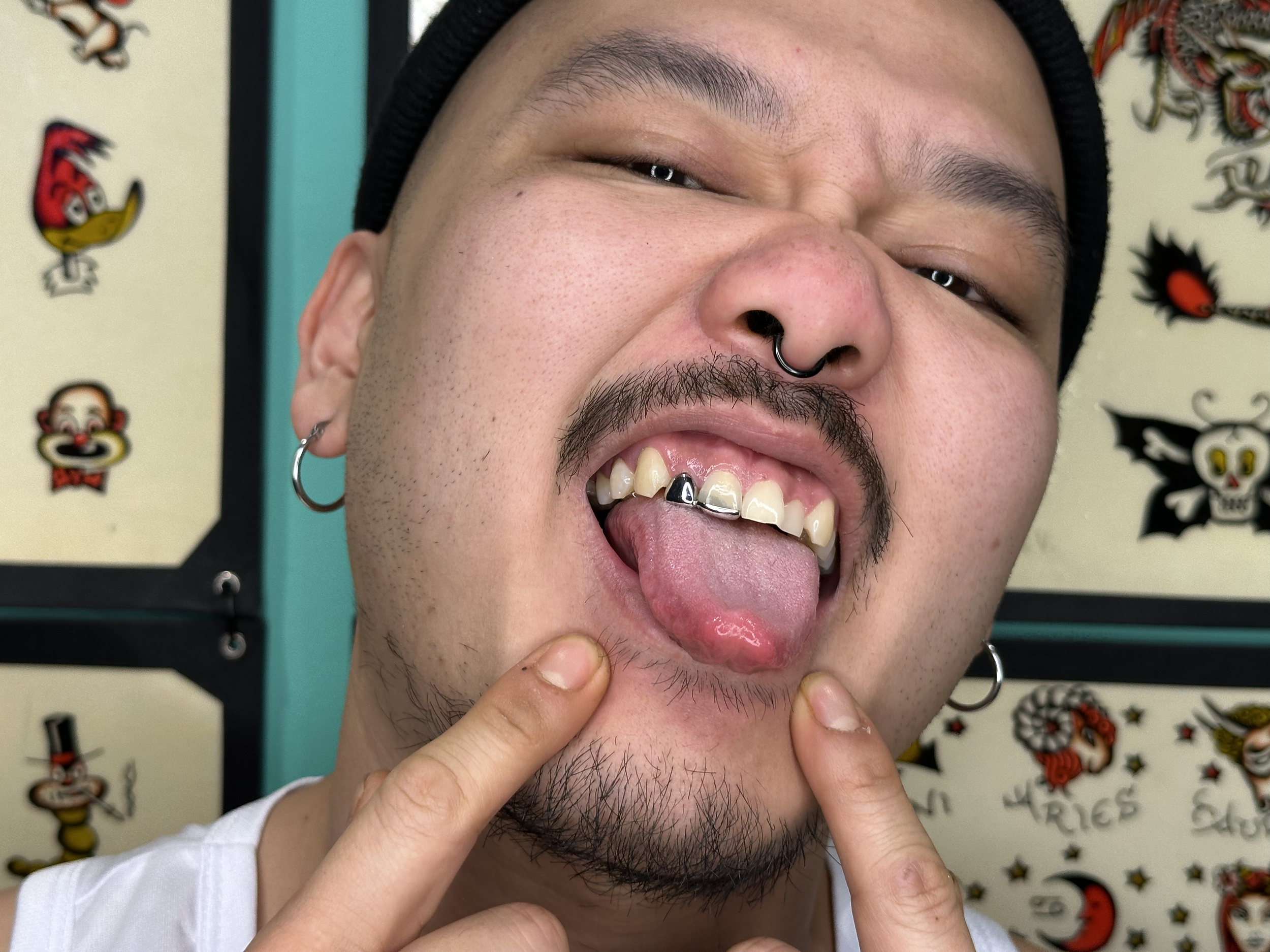 A person with facial piercings, including a septum ring and earrings, showing their teeth and sticking out their tongue while pointing to their chin.
