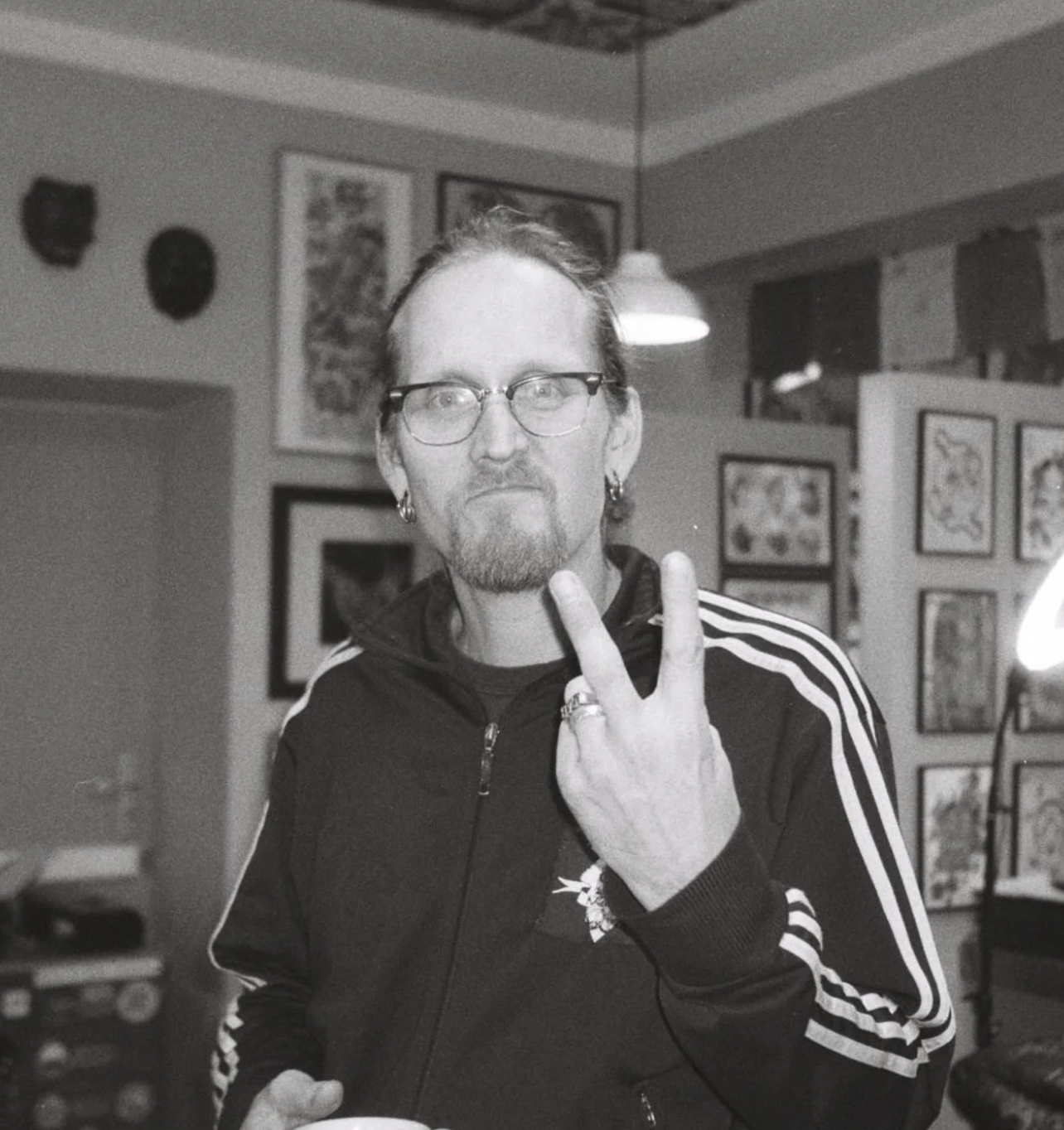 A man with glasses, earrings, and a beard making a peace sign with his fingers in an indoor space decorated with framed pictures and masks.