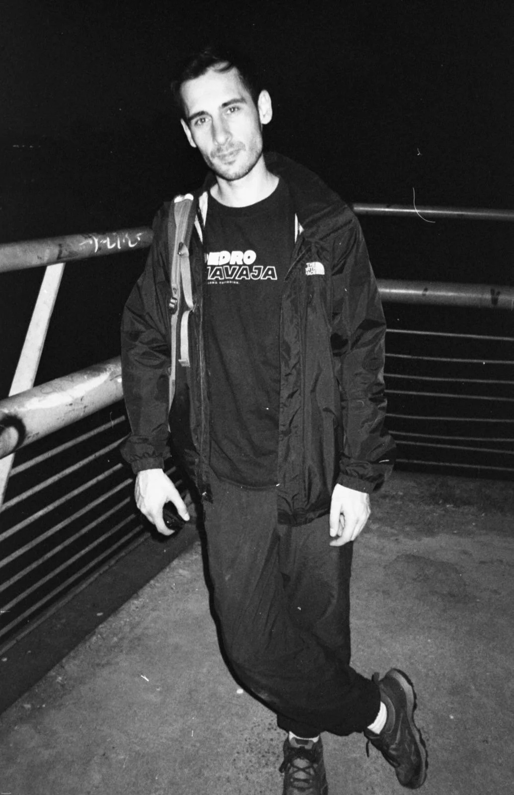 A young man standing on a bridge at night, wearing a black jacket and pants, with a backpack, holding an object in his right hand, and looking at the camera.