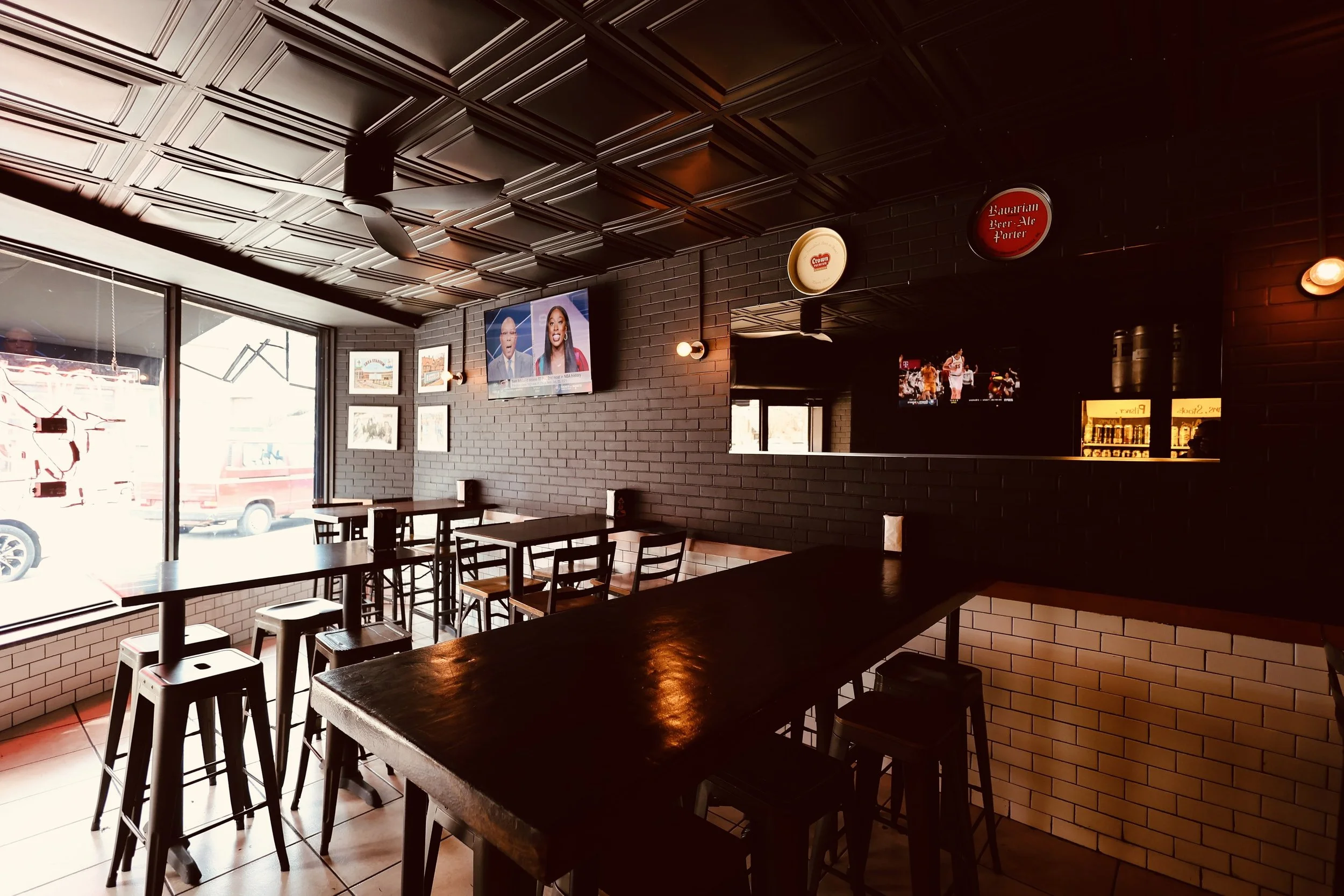 Interior of a modern restaurant or bar with black brick walls, dark wood tables, high chairs, and multiple wall-mounted televisions showing sports and news. Large windows let in natural light, and the ceiling has decorative panels and a ceiling fan.