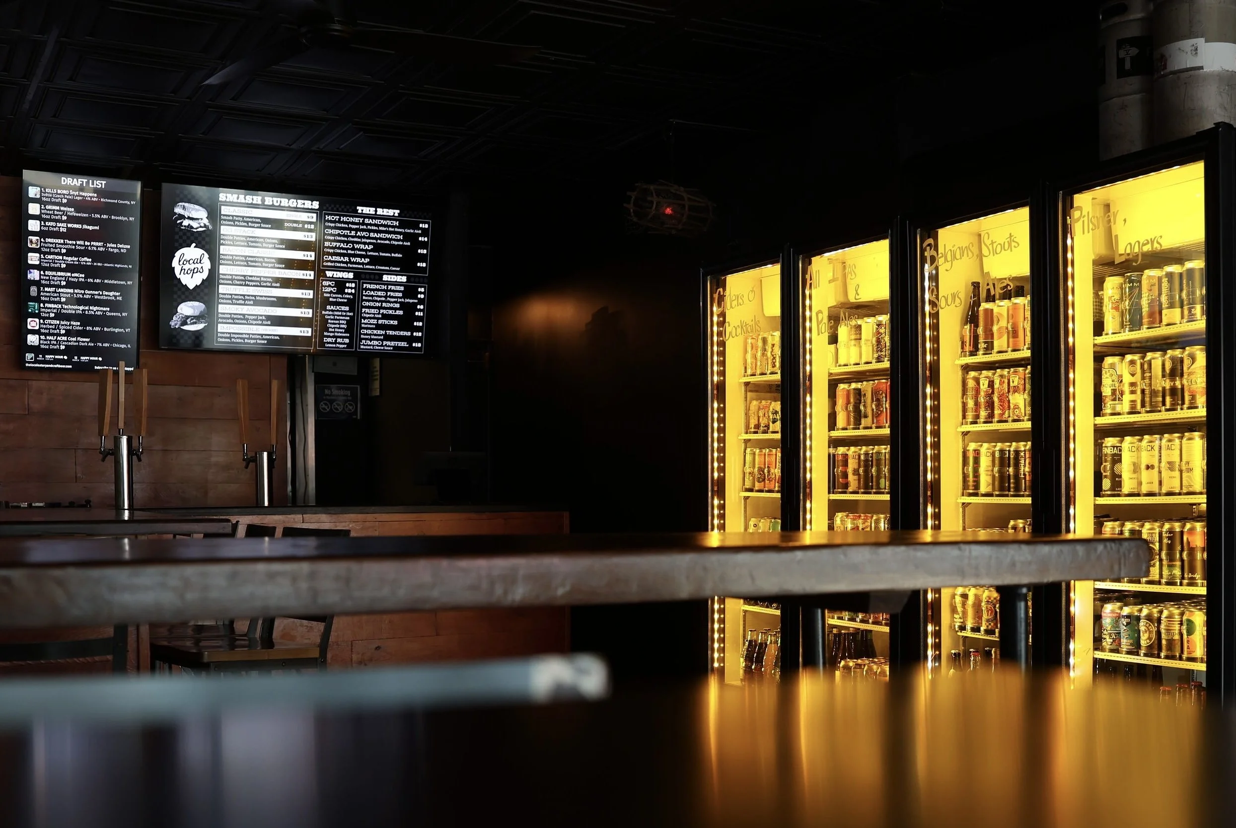 A bar or restaurant counter with two illuminated refrigerators filled with beverages, and digital menu screens displaying food and drink options against a dark background.