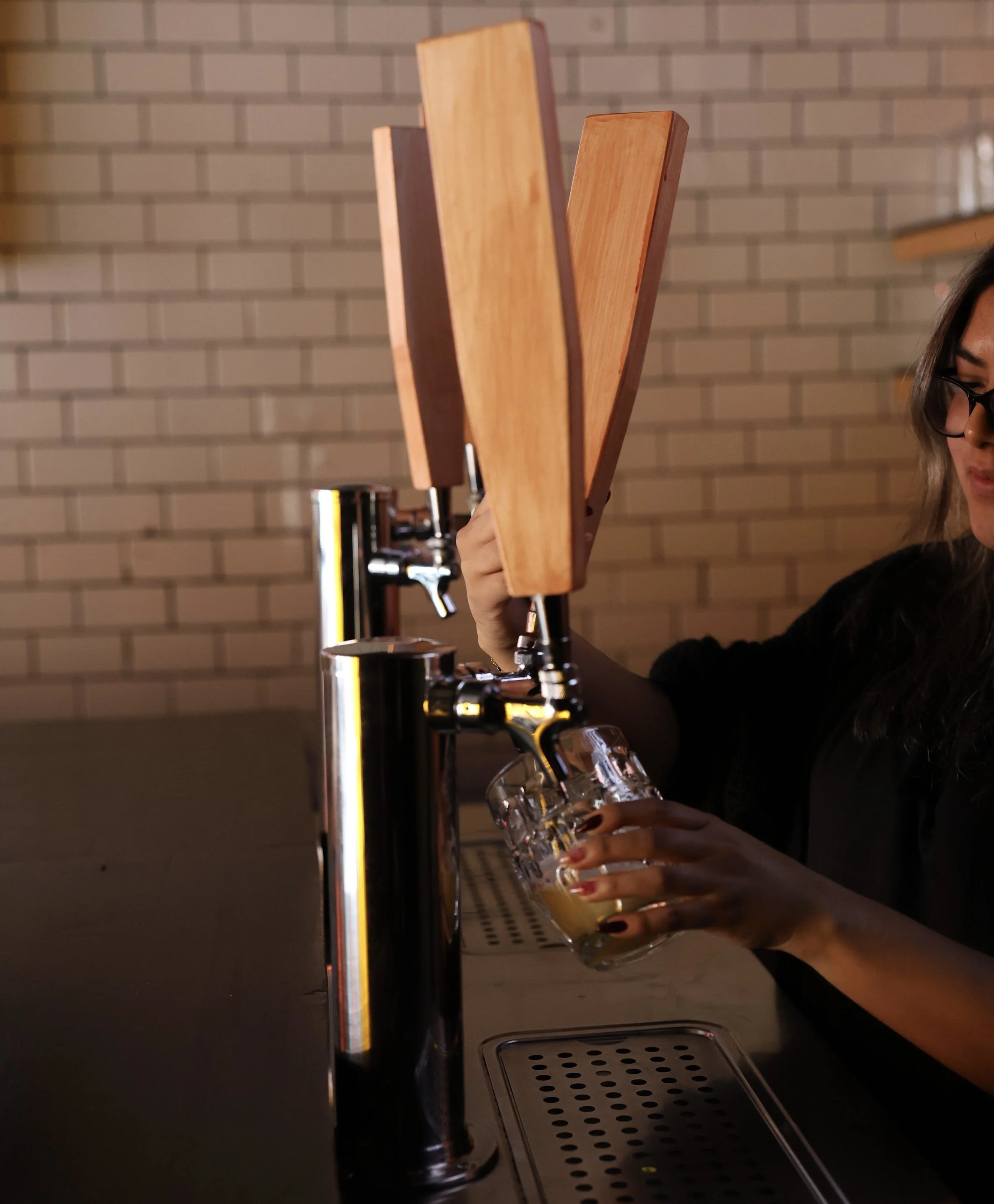 A person holding a glass of beer at a bar, with a row of wooden beer taps and a metal drain on the bar counter, and a brick wall in the background.