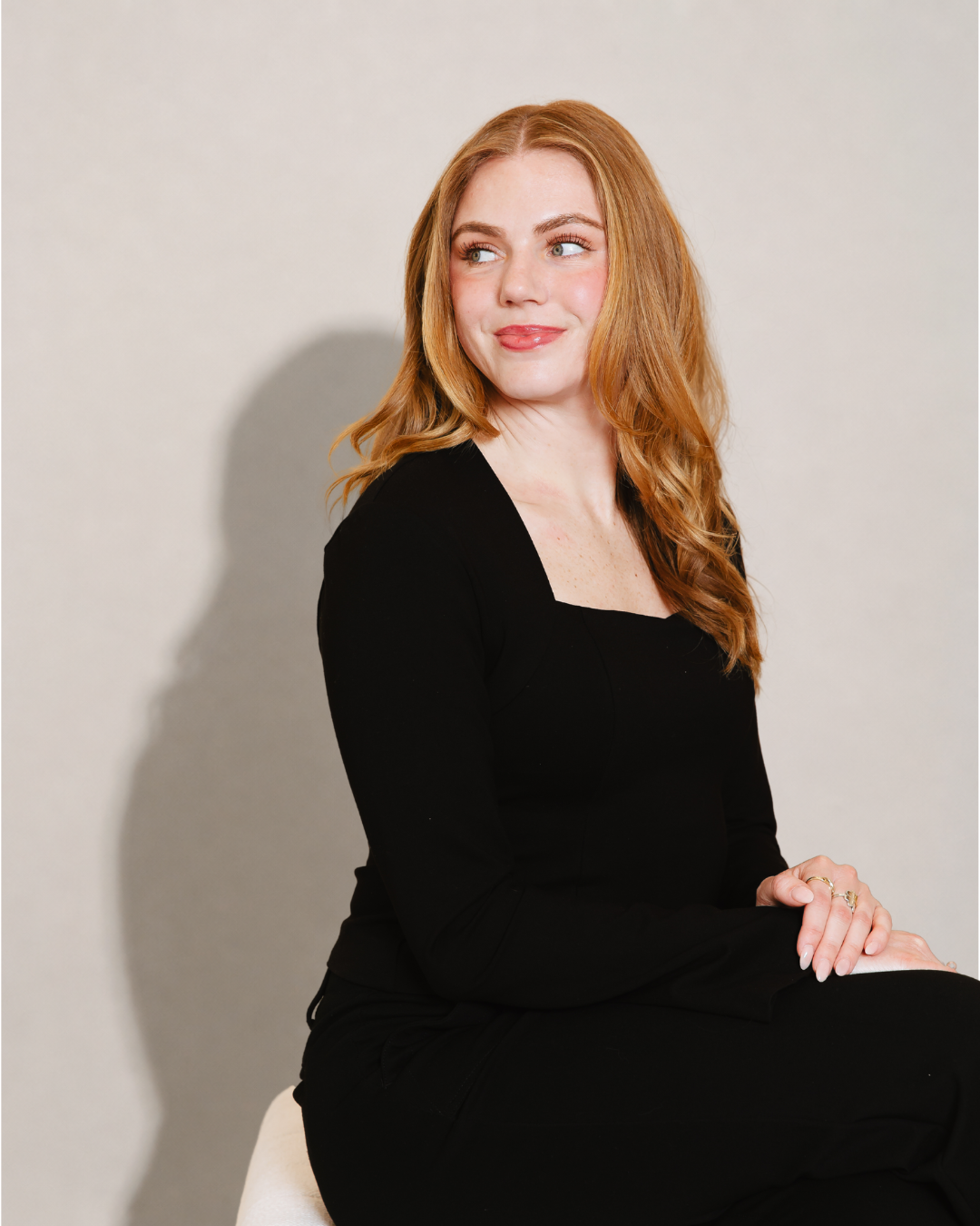 A red-haired woman in black clothing sitting on a white stool against a neutral background, looking to her right with a slight smile.