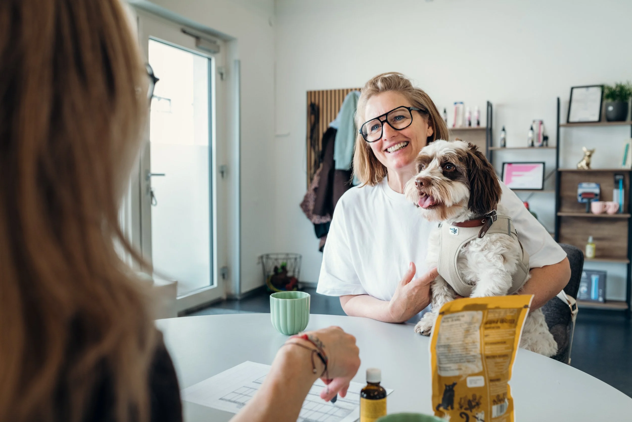 Eine glückliche Frau mit Brille hält einen kleinen Hund im Arm, während sie mit einer anderen Person im Gespräch ist. Der Hund trägt eine Weste und leckt die Zunge heraus. Der Raum ist hell und modern eingerichtet, mit Regalen im Hintergrund.