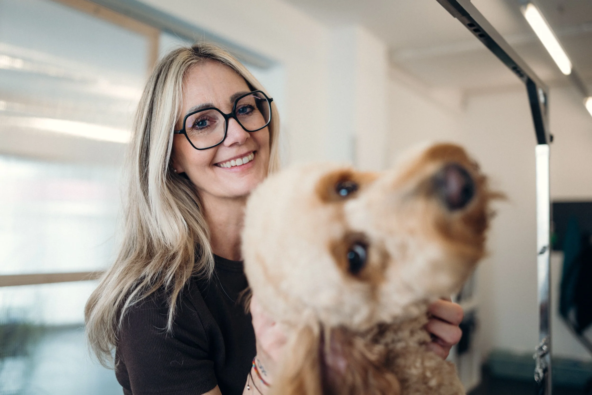 Eine lächelnde Frau mit langen blondem Haar und Brille hält einen Hund in einem Raum, wahrscheinlich Tierpflege oder Tierarzt.