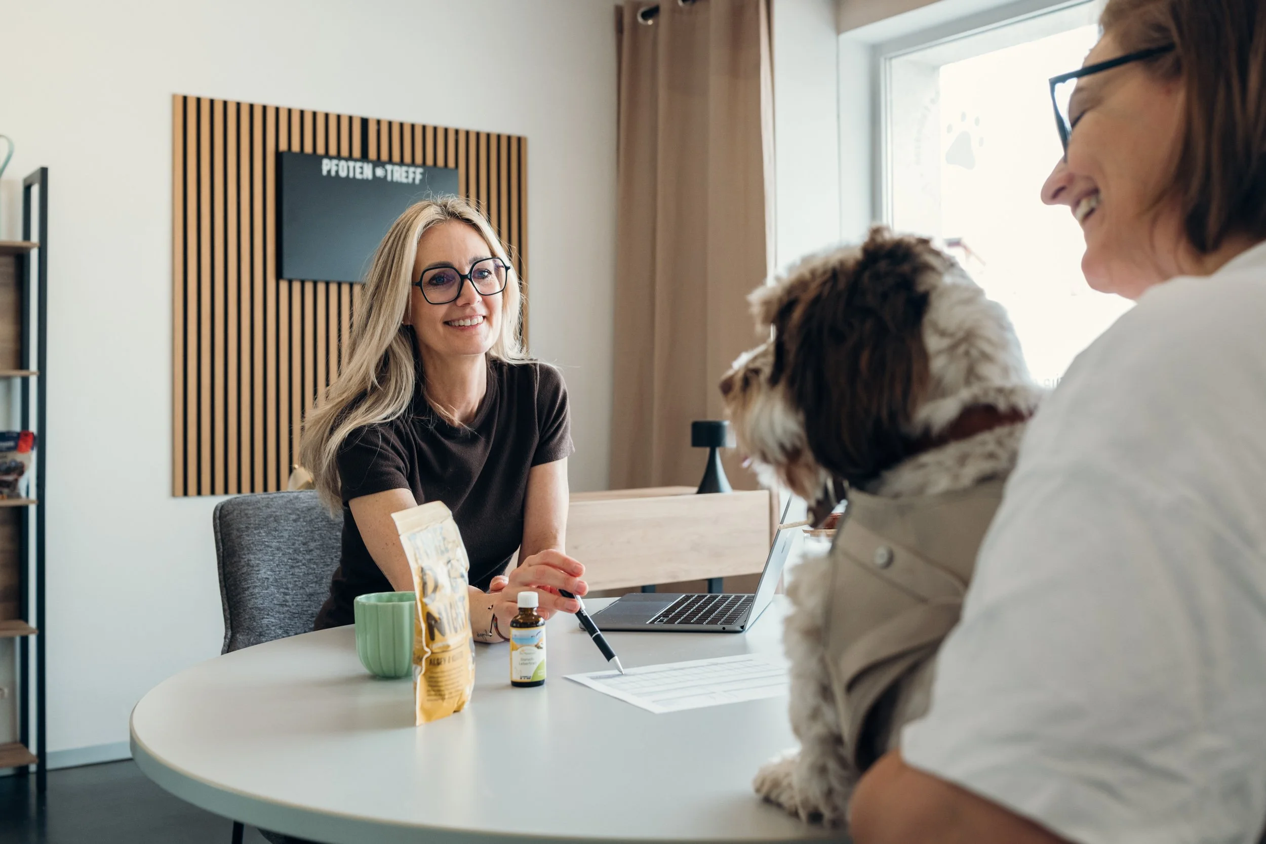 Eine blonde Frau mit Brille sitzt an einem Tisch vor einem Laptop und spricht mit einer anderen Frau, die einen Hund hält. Auf dem Tisch sind Medikamente, eine Tasse und Snacks, im Hintergrund eine Wand mit einem schwarzen Brett.
