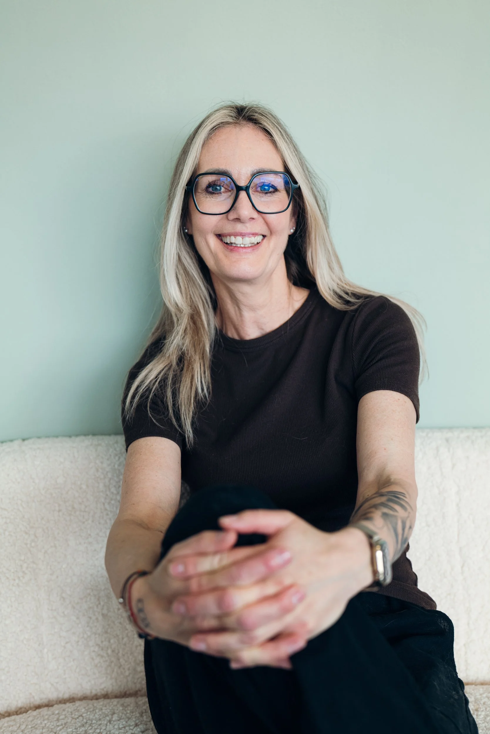 Eine lächelnde Frau mit Brille und langem blonden Haar sitzt auf einem Sofa, hält ein Smartphone in den Händen und trägt ein schwarzes T-Shirt.