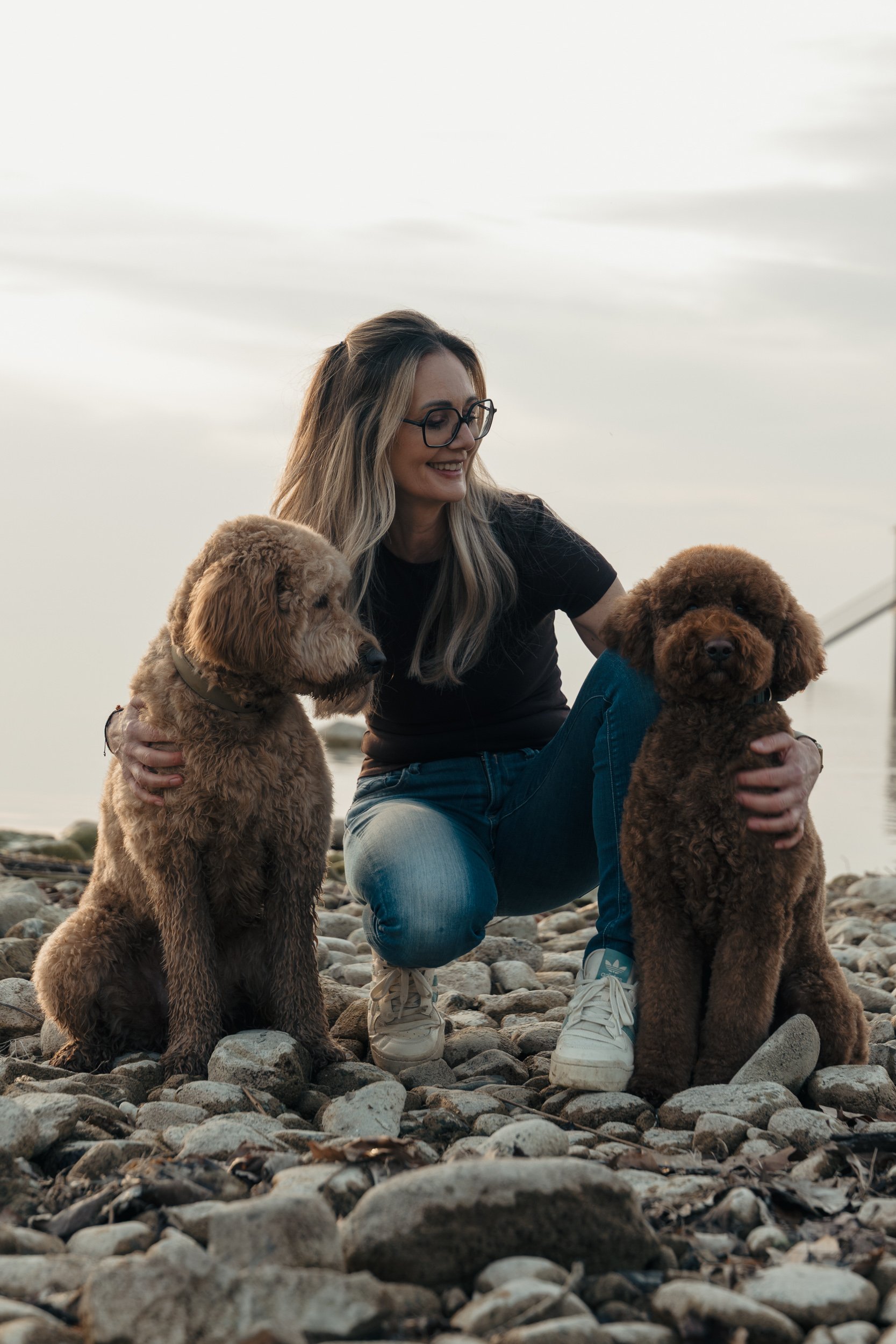 Junge Frau mit langen blonden Haaren und Brille sitzt auf einem Steinstrand, umgeben von zwei braunen Pudeldachshunden, bei bewölktem Himmel.