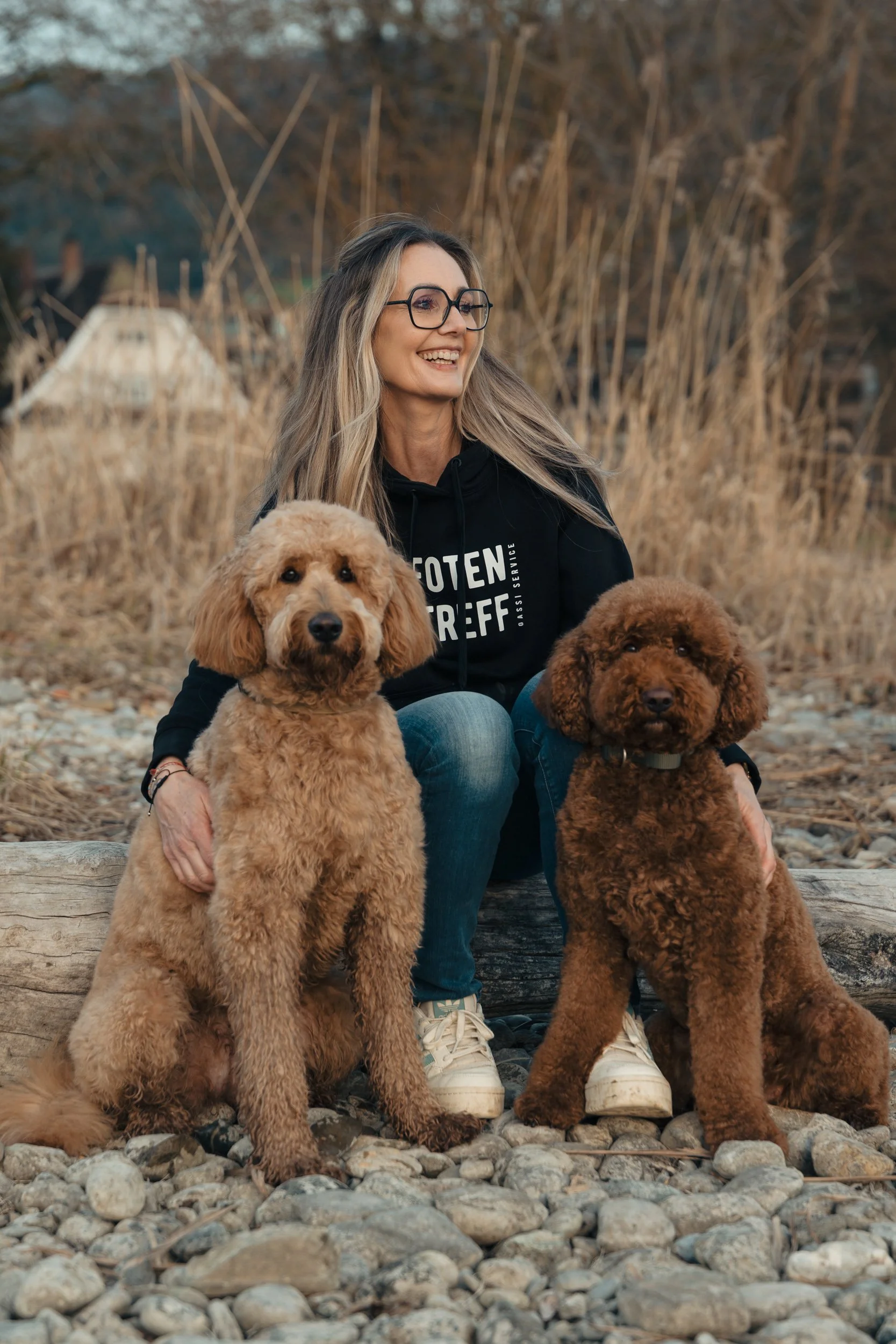 Eine lachende Frau mit Brille sitzt auf einem Baumstamm am Bodensee in Vorarlberg und umarmt zwei braune Labradoodle-Hunde mitten in einer natürlichen Umgebung mit Kieselsteinen, trockenen Pflanzen und Bäumen im Hintergrund.