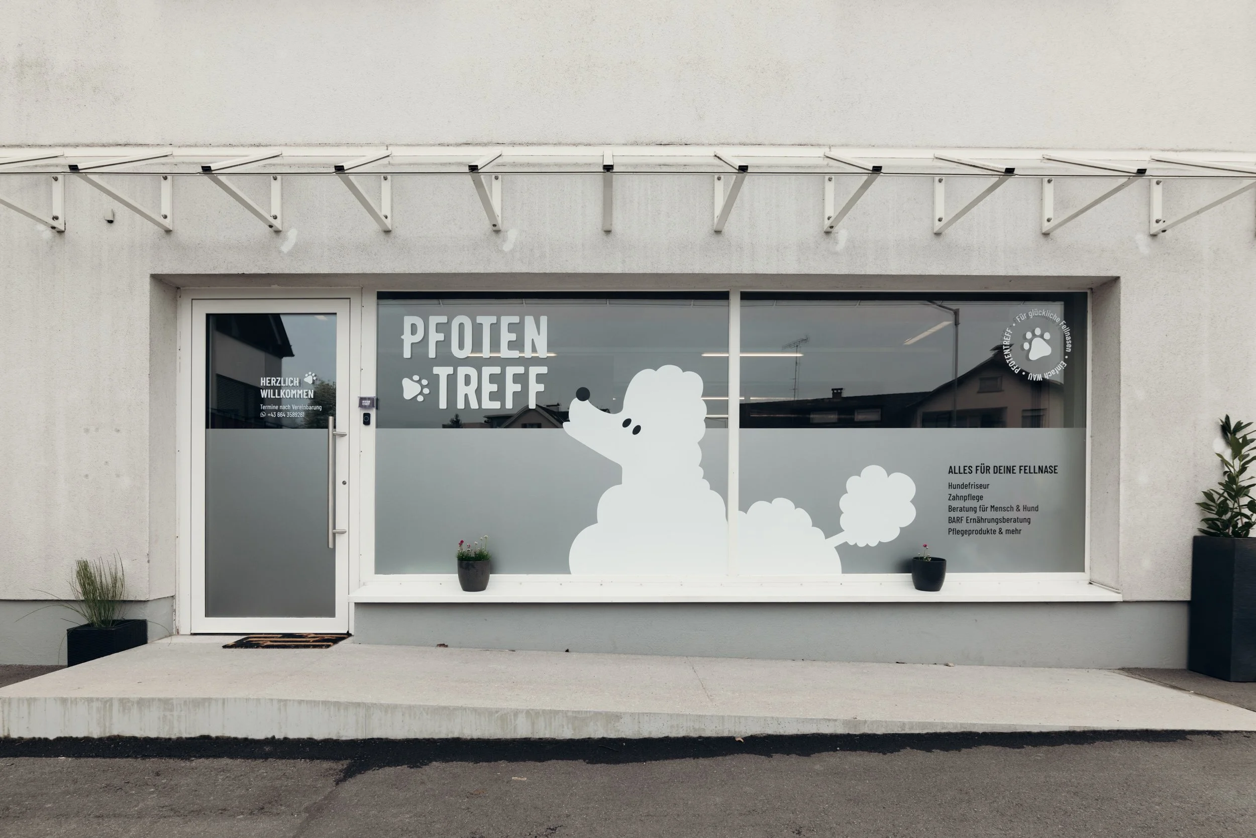 Das Bild zeigt das Schaufenster eines Hundesalons namens "Pfoten Treff". Das Schaufenster ist verziert mit einem großen weißen Hundekopf und Pfotenabdrücken. Es gibt zwei Pflanzkübel mit Pflanzen vor dem Fenster. An der Tür ist ein Willkommensschild mit Angaben zu Terminen und Kontaktinformationen.