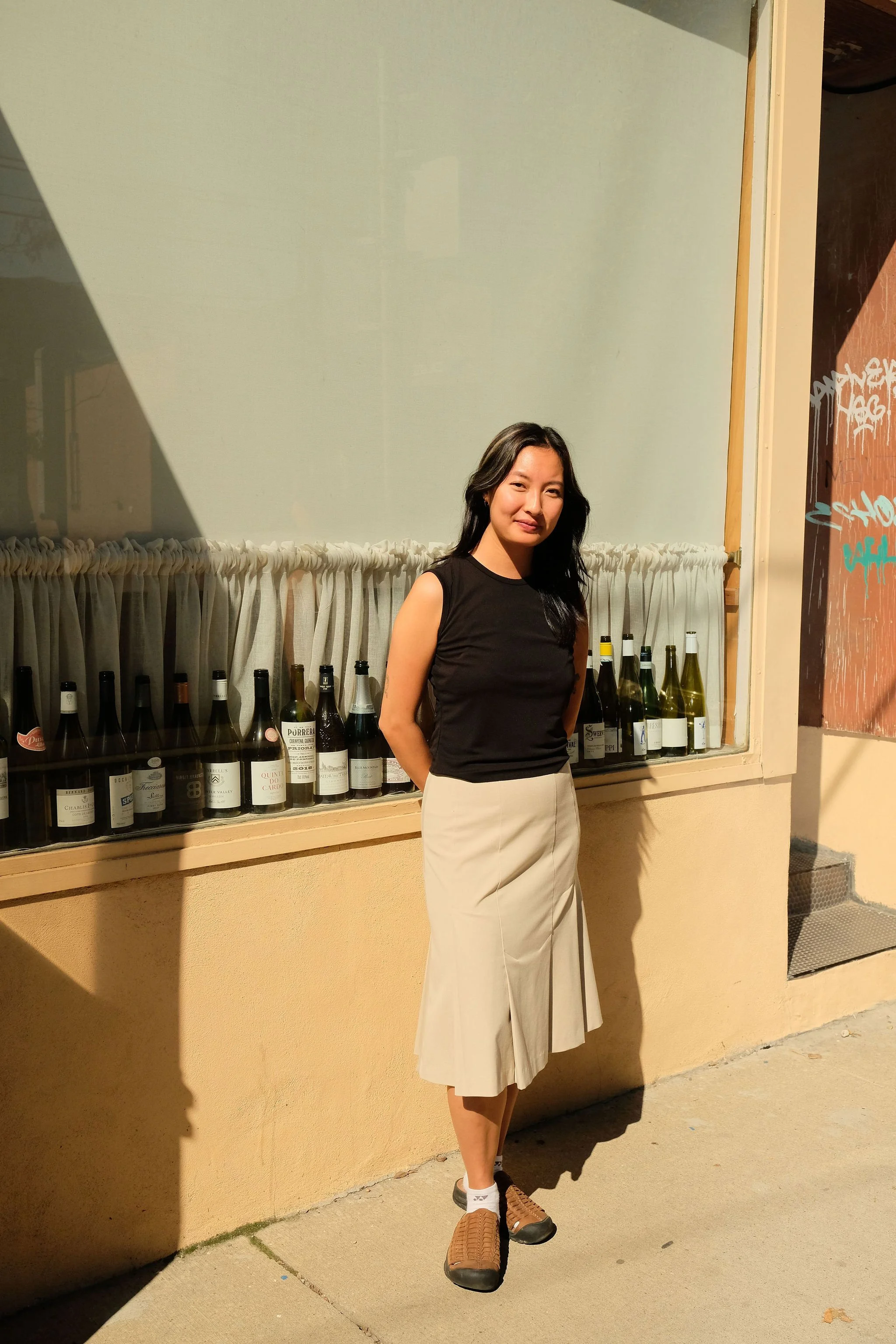A woman standing outside near a glass window with wine bottles, wearing a black sleeveless top, beige skirt, and sneakers, with sunlight casting shadows.