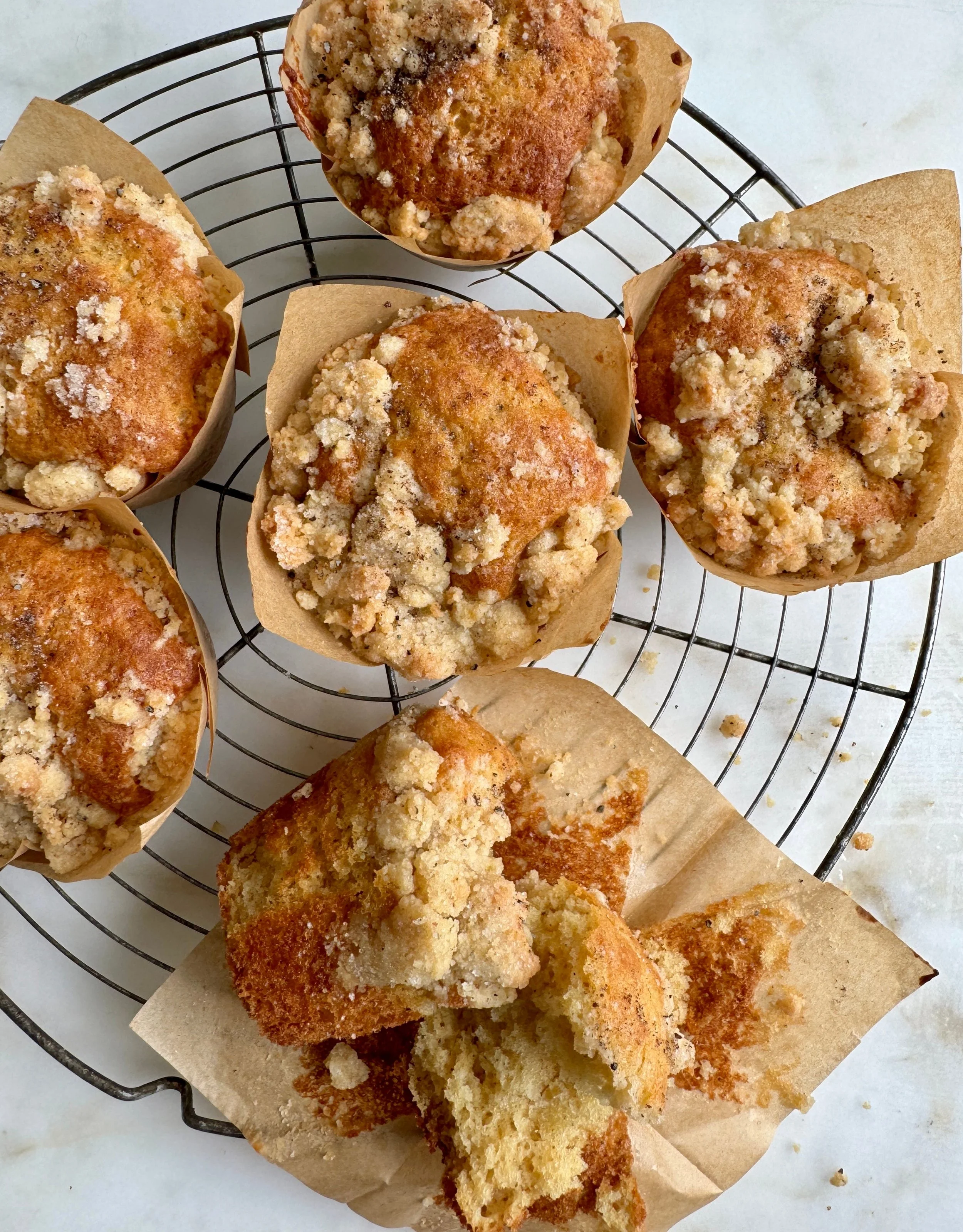 Freshly baked crumb-topped muffins on a wire cooling rack, with some broken pieces and crumbs on parchment paper.