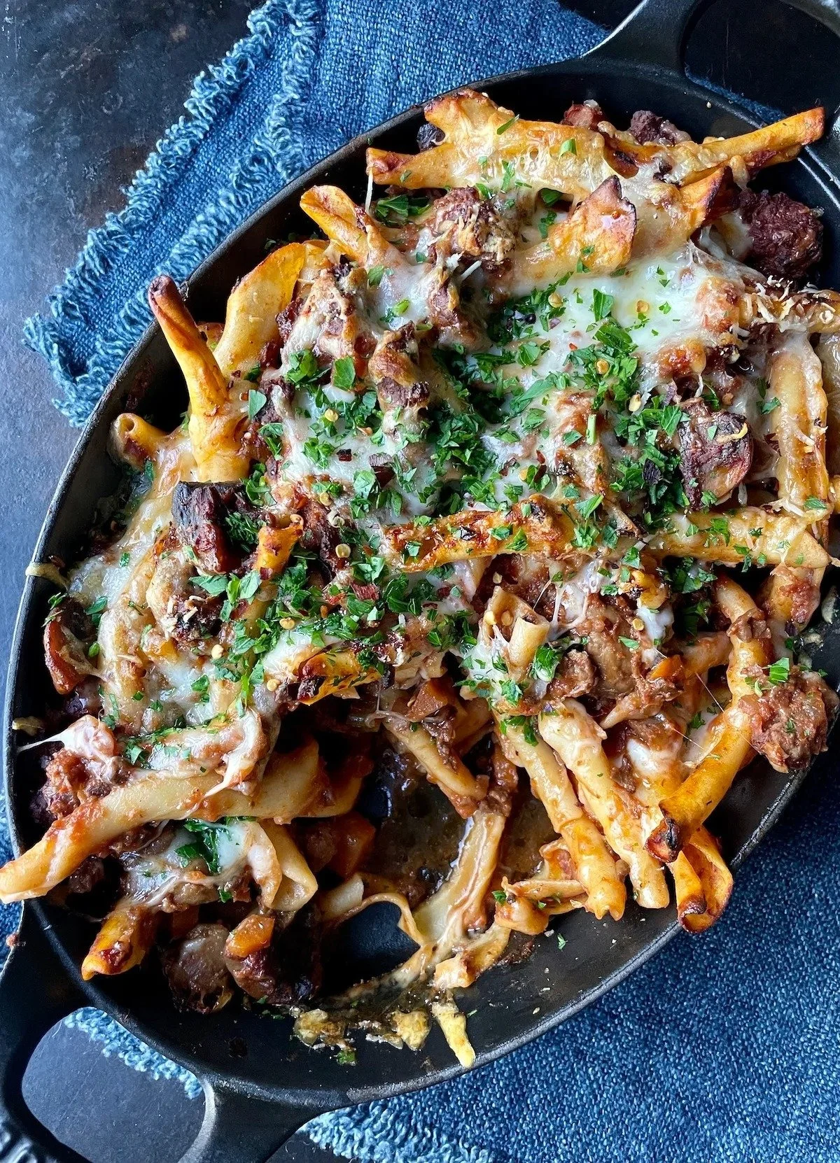 Baked pasta casserole topped with cheese, chopped parsley, and ground meat, served on a black skillet.