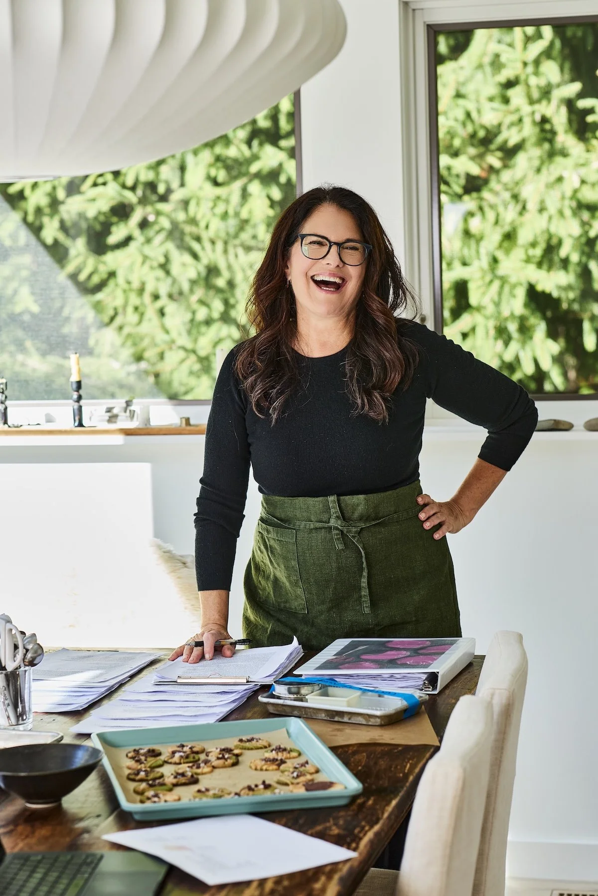 Woman (author Susan Spungen) with long brown hair, glasses, wearing a black top and green apron, smiling and standing in a well-lit modern kitchen with large windows and green foliage outside.