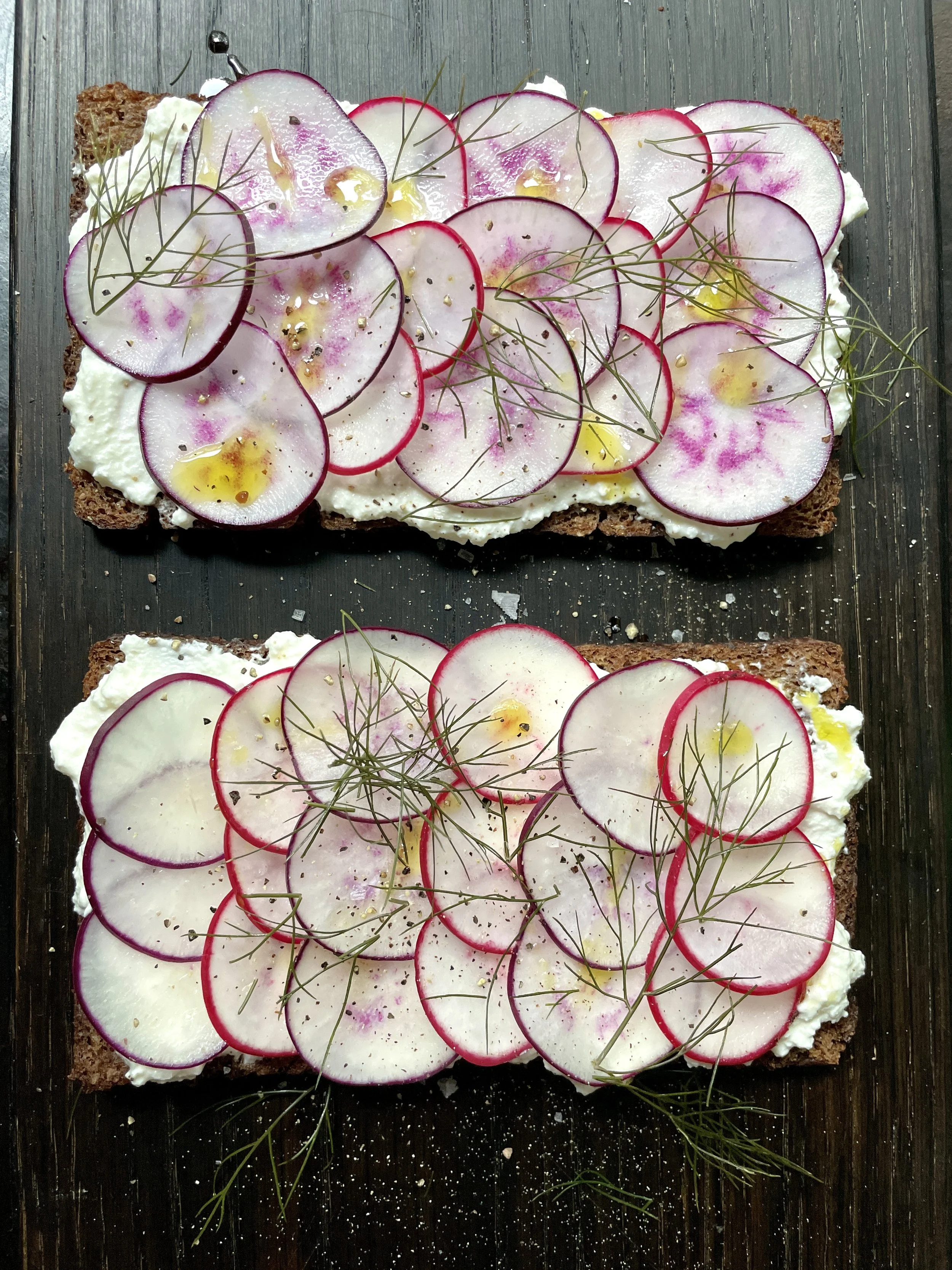 Rectangular slices of bread topped with creamy spread, thinly sliced radishes, sprigs of herbs, and sprinkled with black pepper and gold-colored seasoning on a black surface.