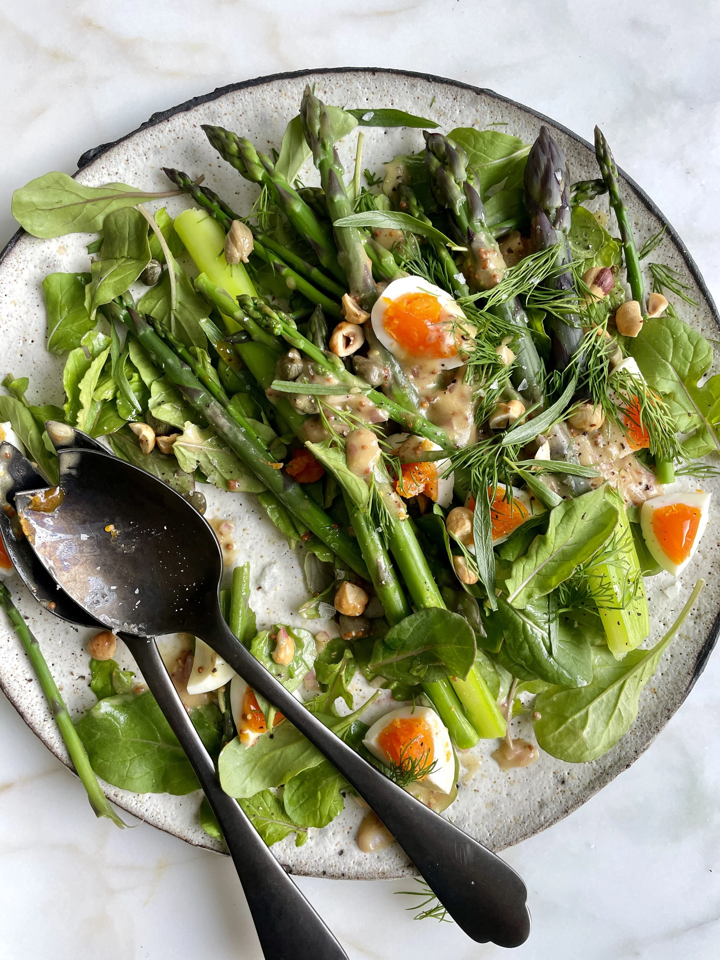 A plate of salad with asparagus, greens, chopped nuts, and halved soft-boiled eggs. Two black serving spoons rest on the plate.