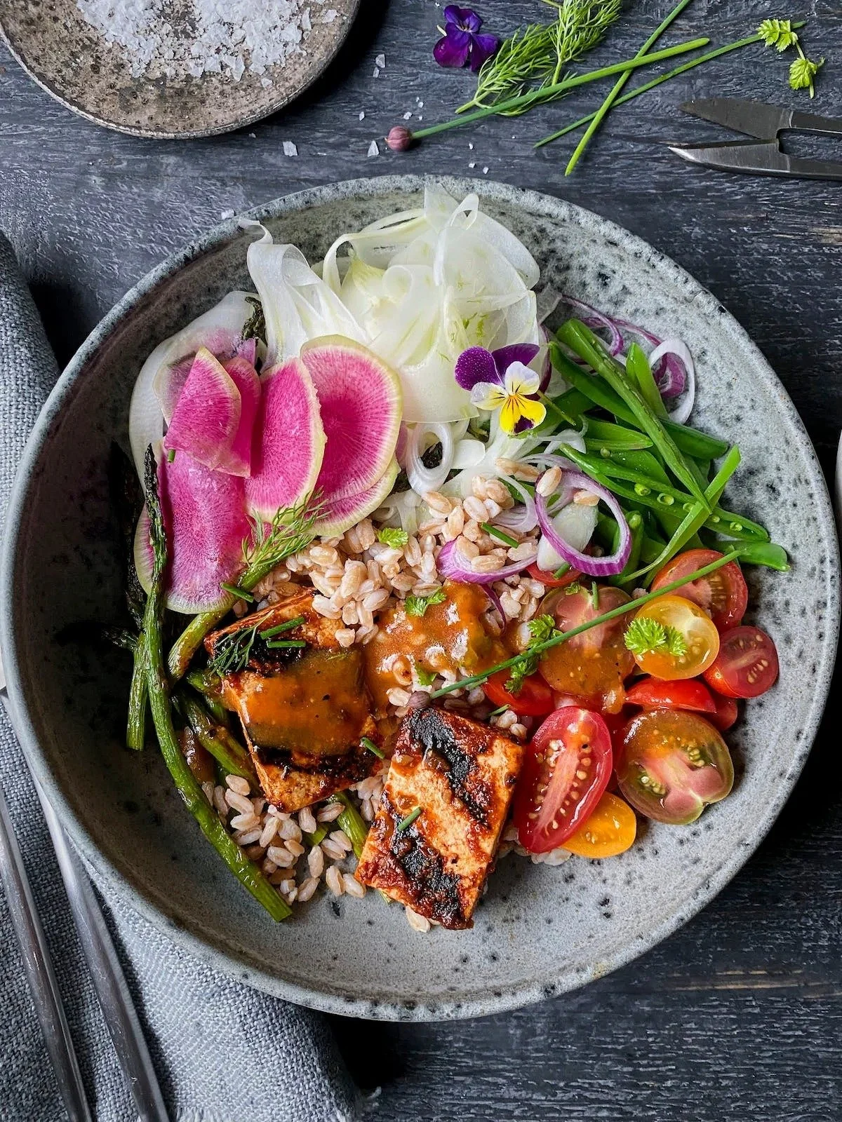 A bowl of colorful salad containing grilled fish pieces, cherry tomatoes, sliced radishes, onion slices, chopped grains, and fresh herbs, garnished with edible flowers, on a rustic dark table.