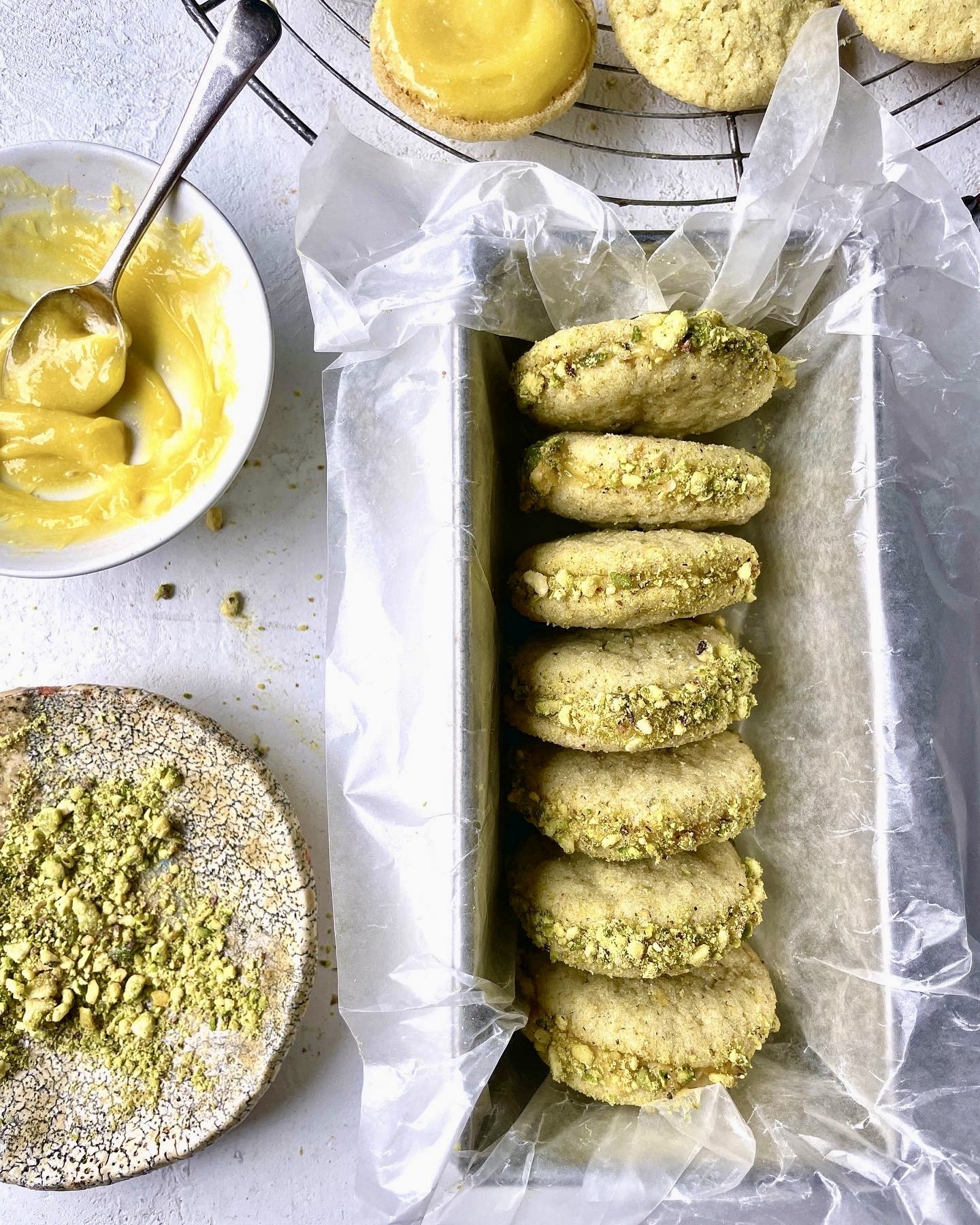 Assorted cookies, some topped with chopped pistachios, on a white surface. A bowl of lemon curd and a wire rack with more cookies are also visible.