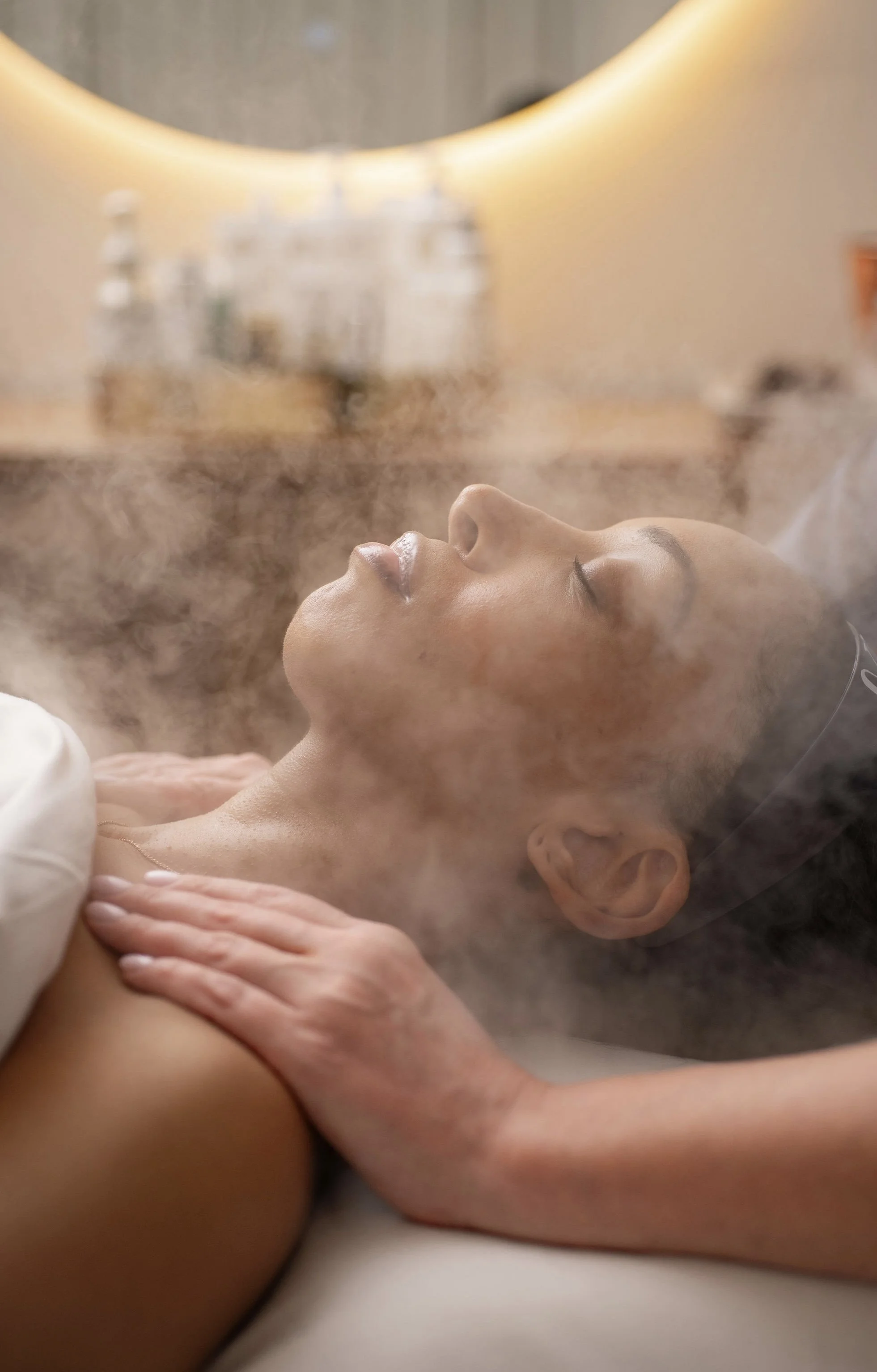A woman lying down during a steam facial treatment, with steam surrounding her face and an aesthetician's hands on her shoulder.