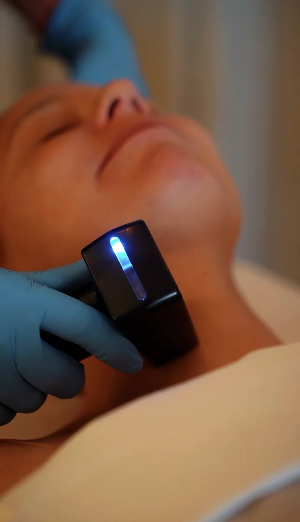 A person receiving a laser skin treatment on their face in a medical setting, with a technician wearing blue gloves holding a handheld device emitting light.